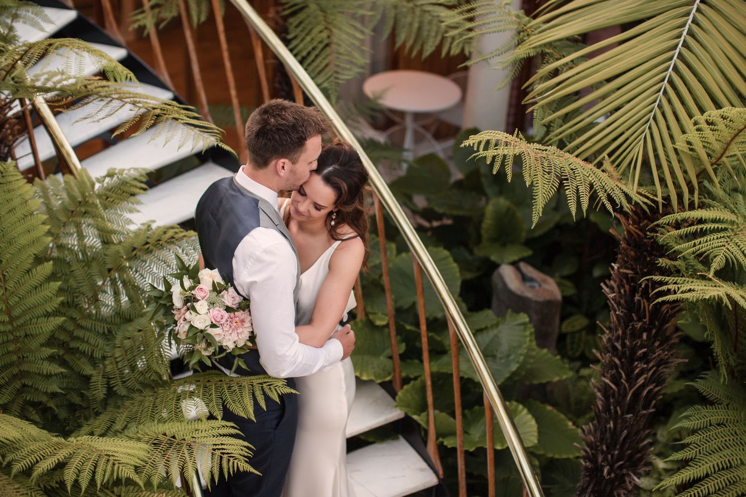 A couple shares a romantic kiss under tropical palm trees during an outdoor wedding ceremony.