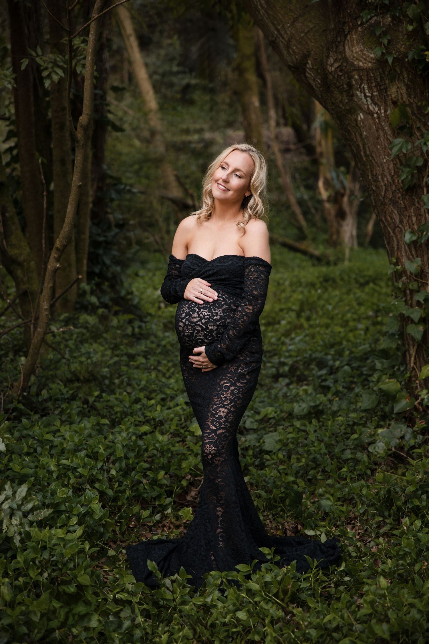 Pregnant woman in black lace off-shoulder gown cradling baby bump in lush green forest setting.