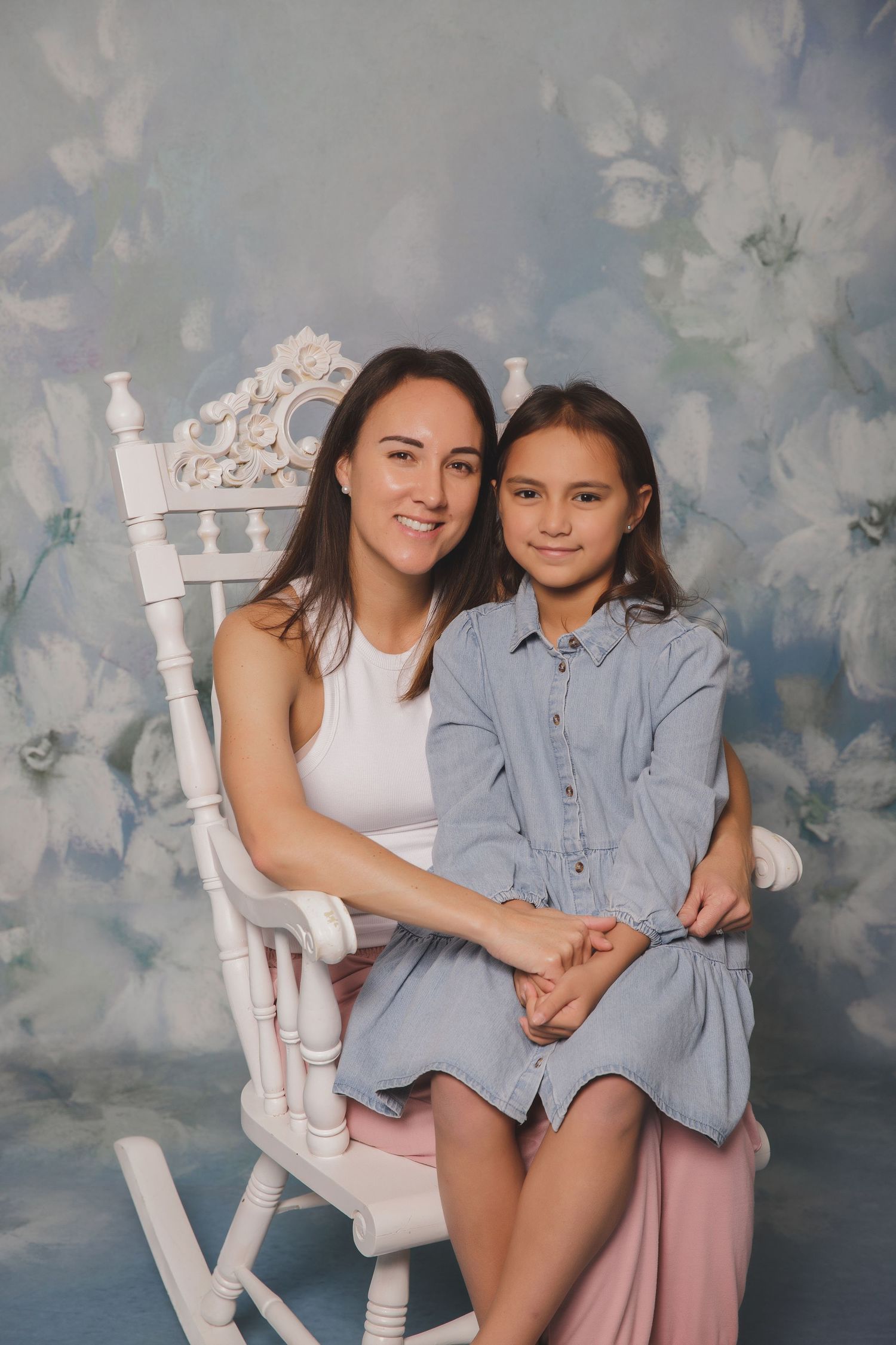 A woman and young girl pose together on a white rocking chair in front of a floral backdrop.