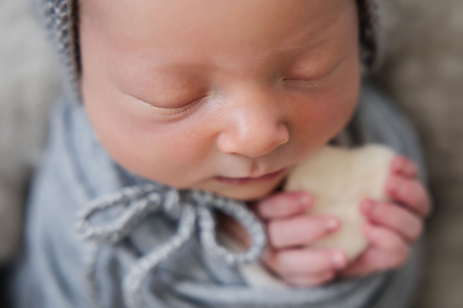 Peaceful newborn baby sleeping wrapped in gray swaddle, wearing knit hat, holding small heart prop in tiny hands.
