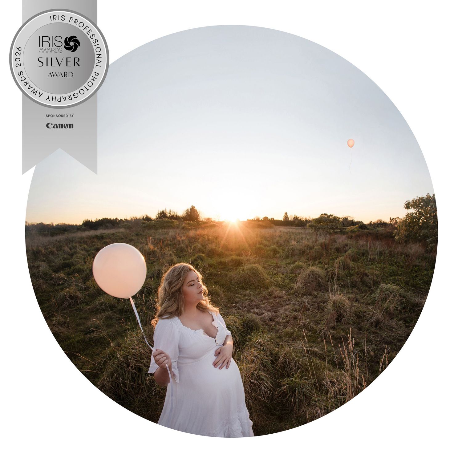 Pregnant woman in white dress holding pink balloon at golden sunset in a field, IRIS Silver Award winning maternity photo.