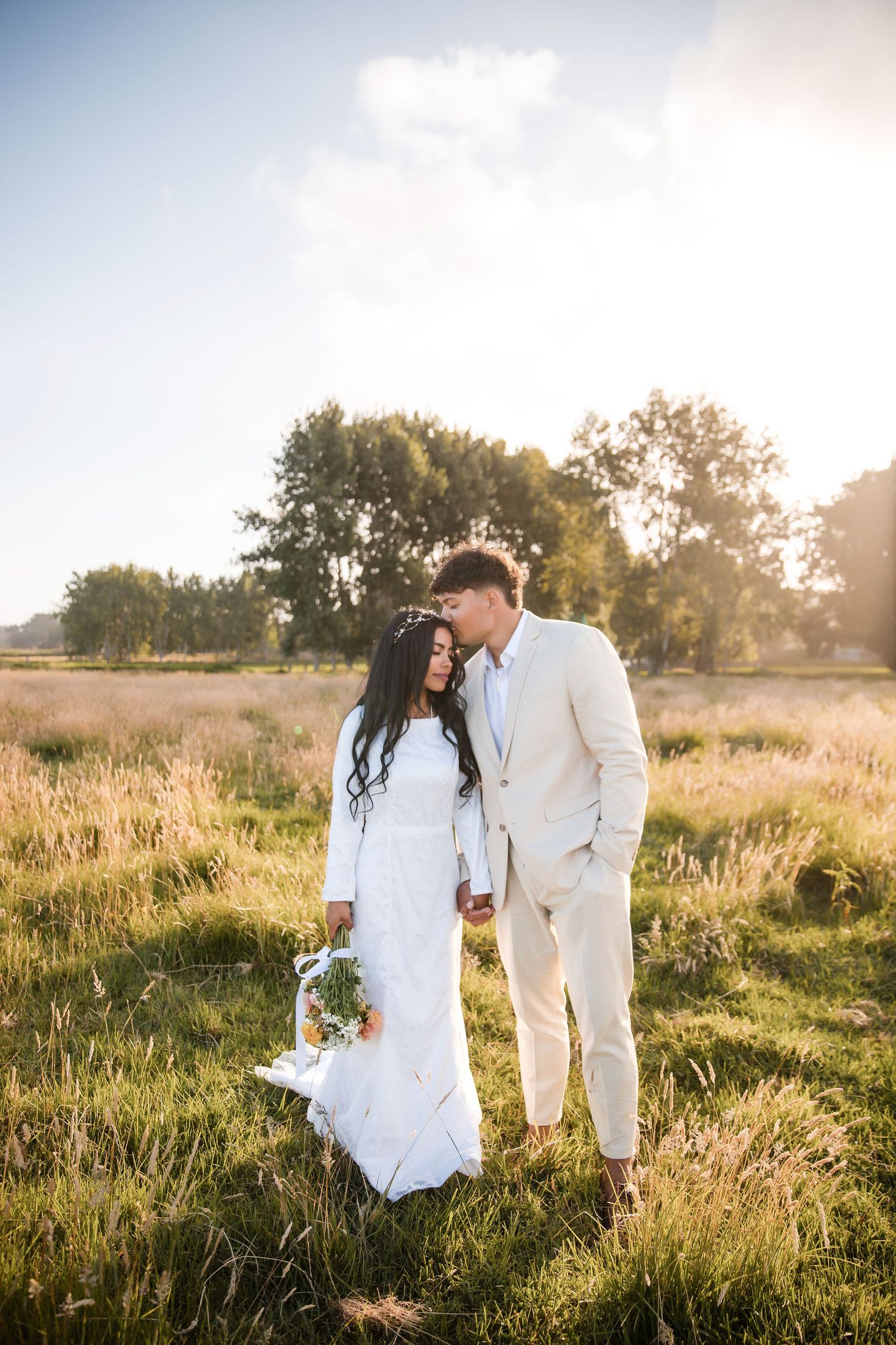 Couple in white attire standing in a sunlit meadow during golden hour photography session.