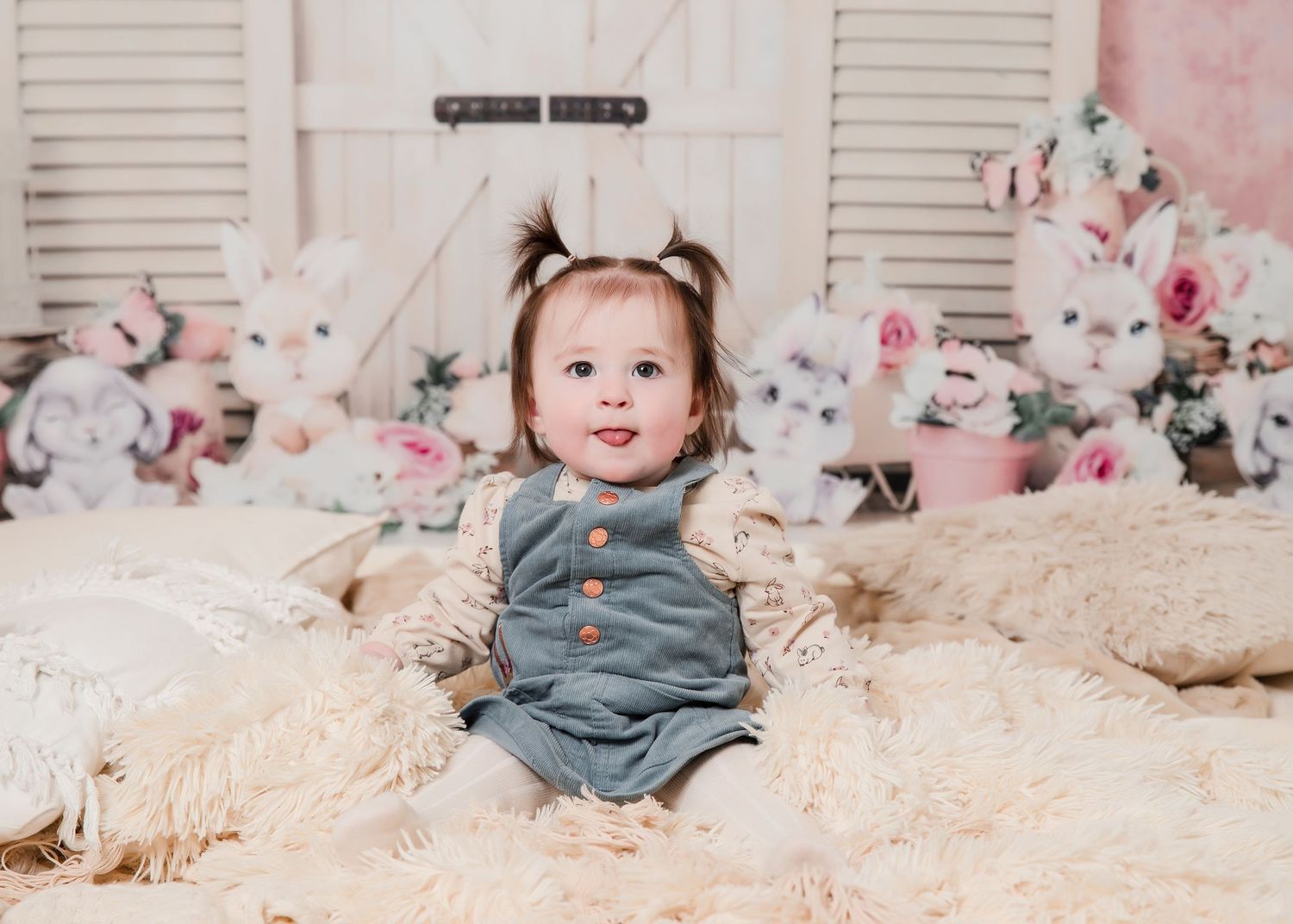 Baby in pink floral dress sits among soft white blankets and teddy bears during a birthday photo shoot.