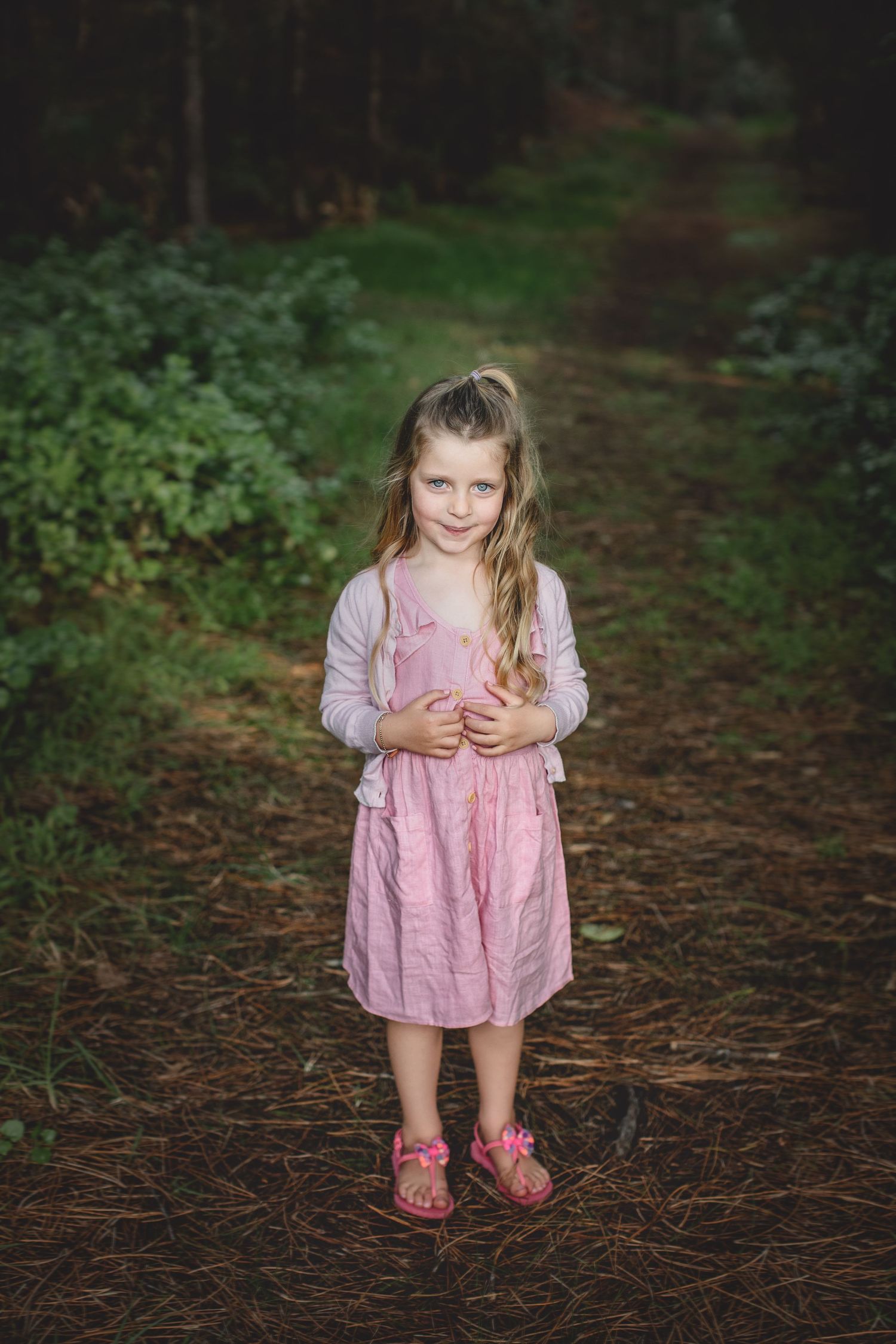 Young girl in pink dress stands on forest path wearing pink sandals.