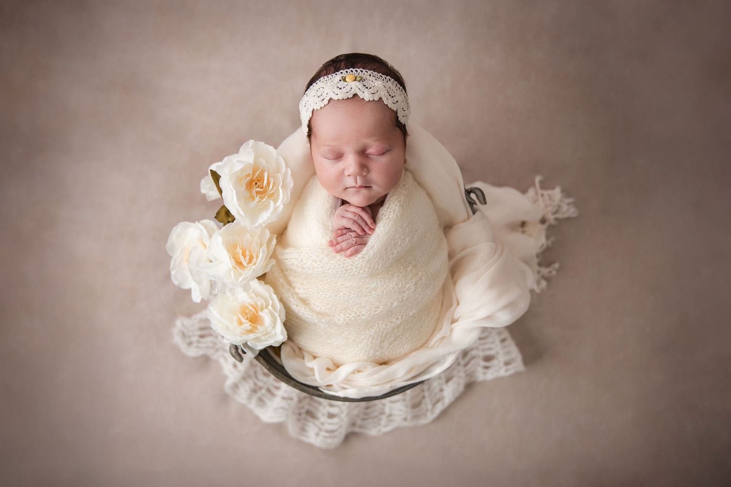 A sleeping newborn baby lies peacefully in a cream-colored wrap on a soft lace blanket in a studio setting.