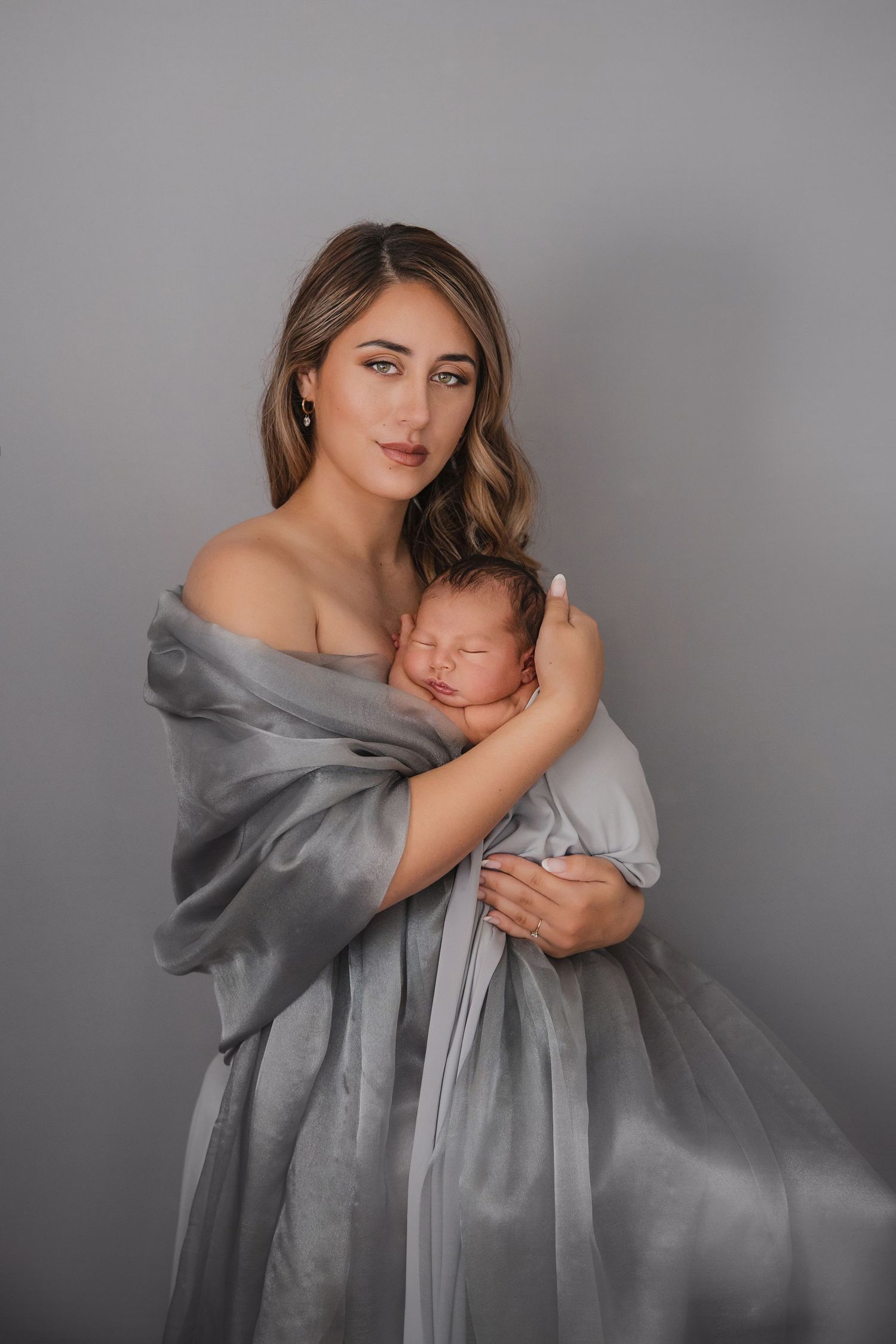A mother wrapped in a gray silk shawl tenderly holds her sleeping newborn baby against a gray studio background.