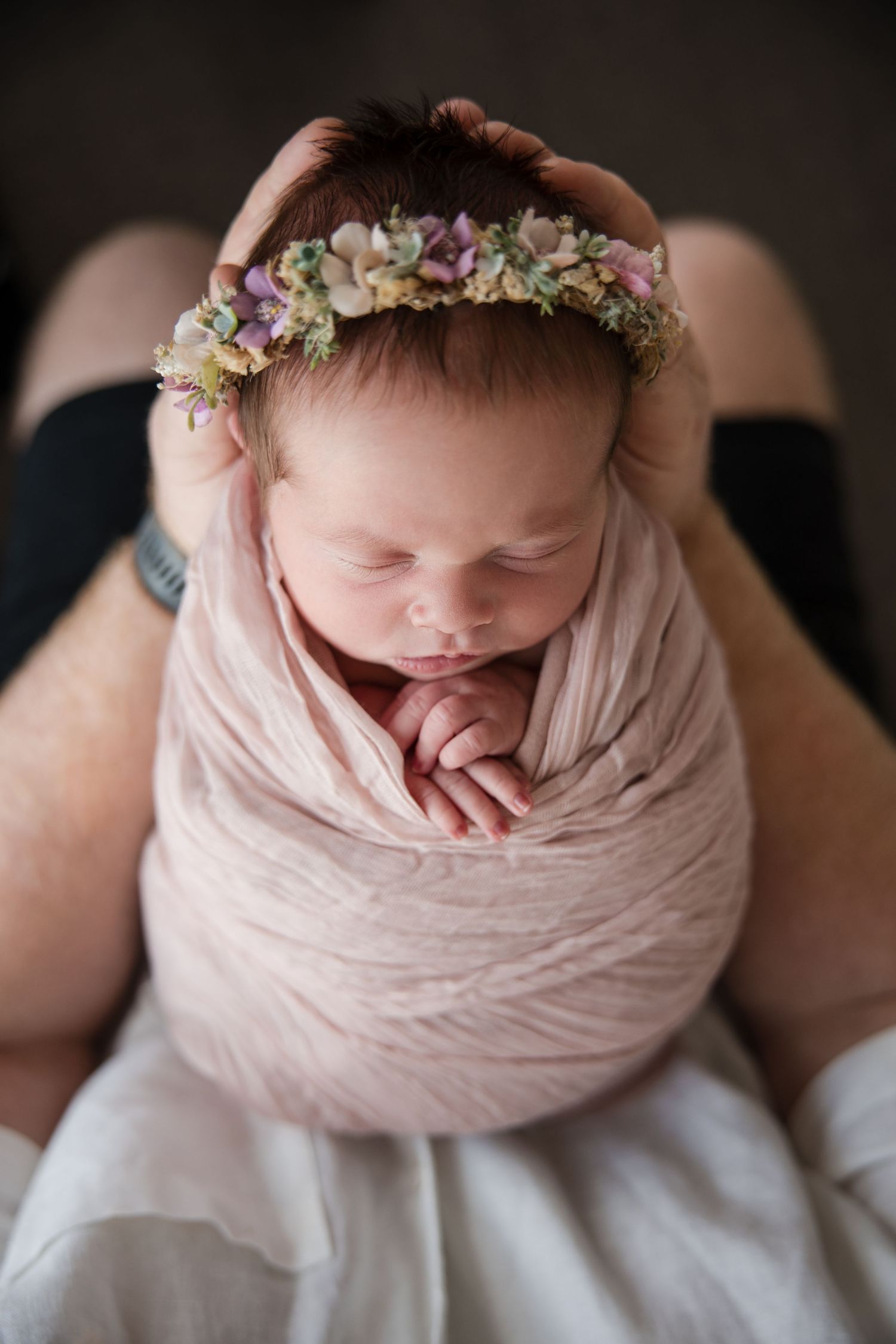 Newborn baby wrapped in soft pink fabric wearing delicate floral crown against dark background.