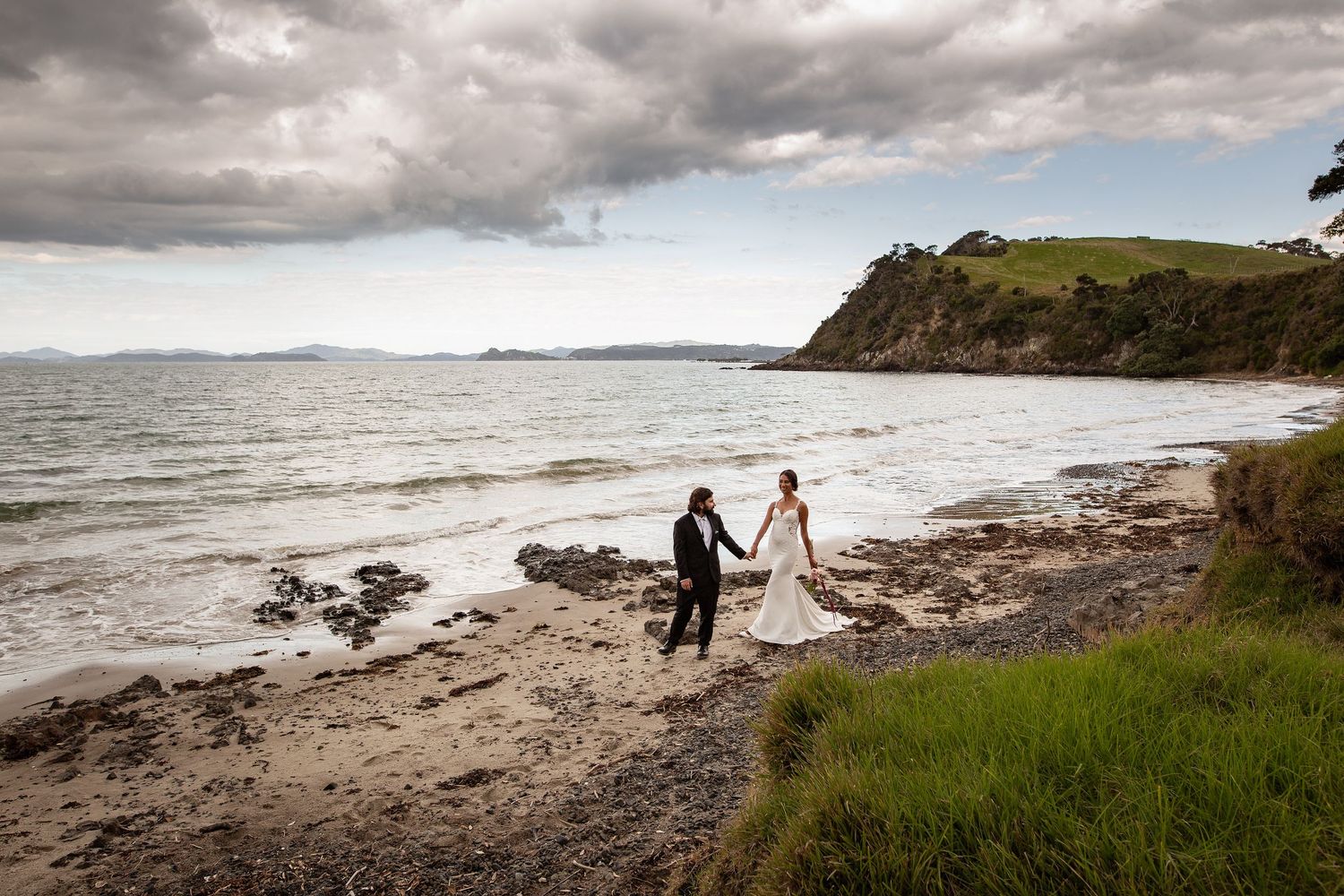 A couple walks hand in hand on a dramatic rocky beach with stormy clouds and cliffs along the New Zealand coastline.
