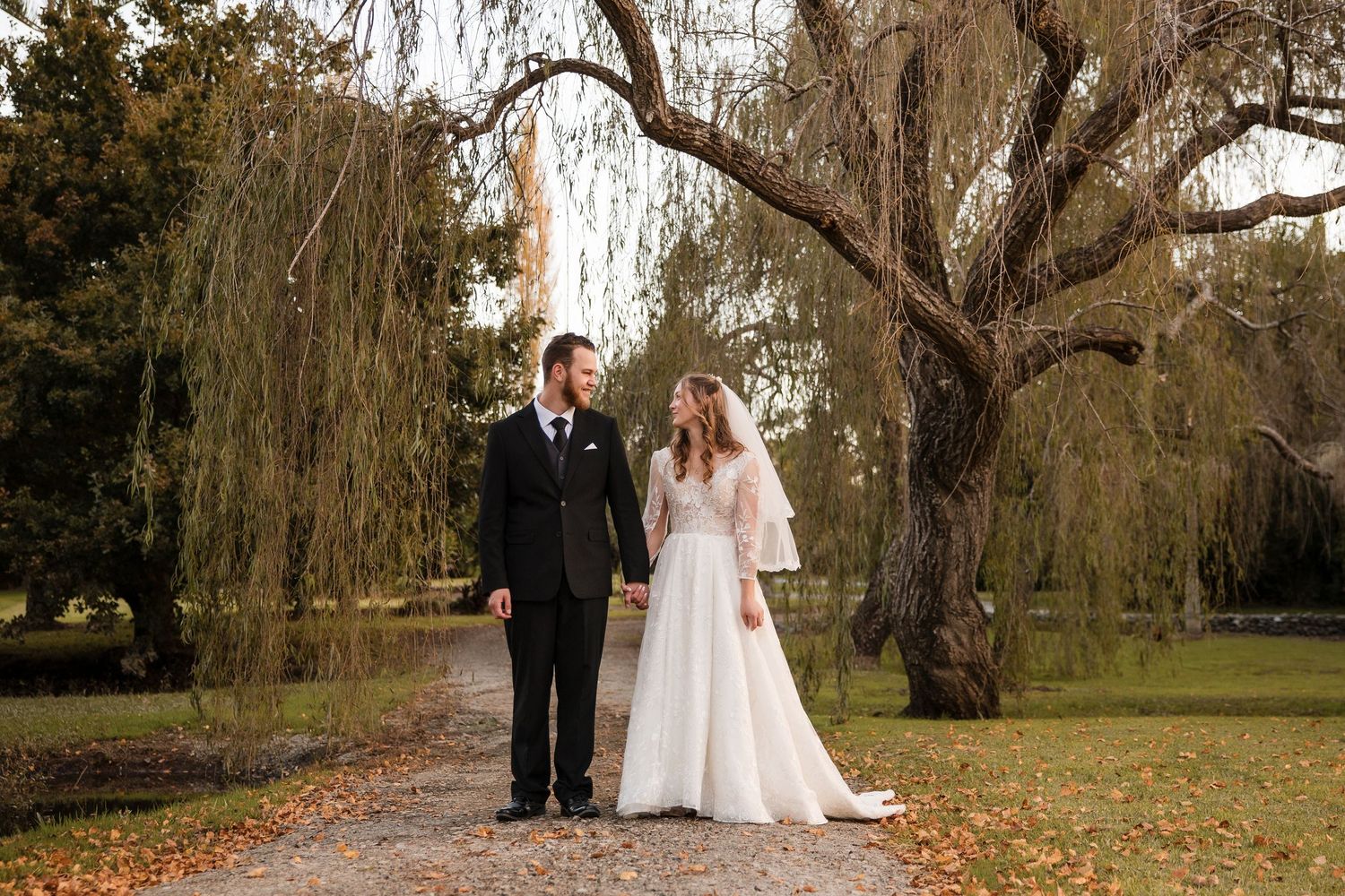 A couple stands together on an autumn day beneath large weeping willow trees in a park setting.