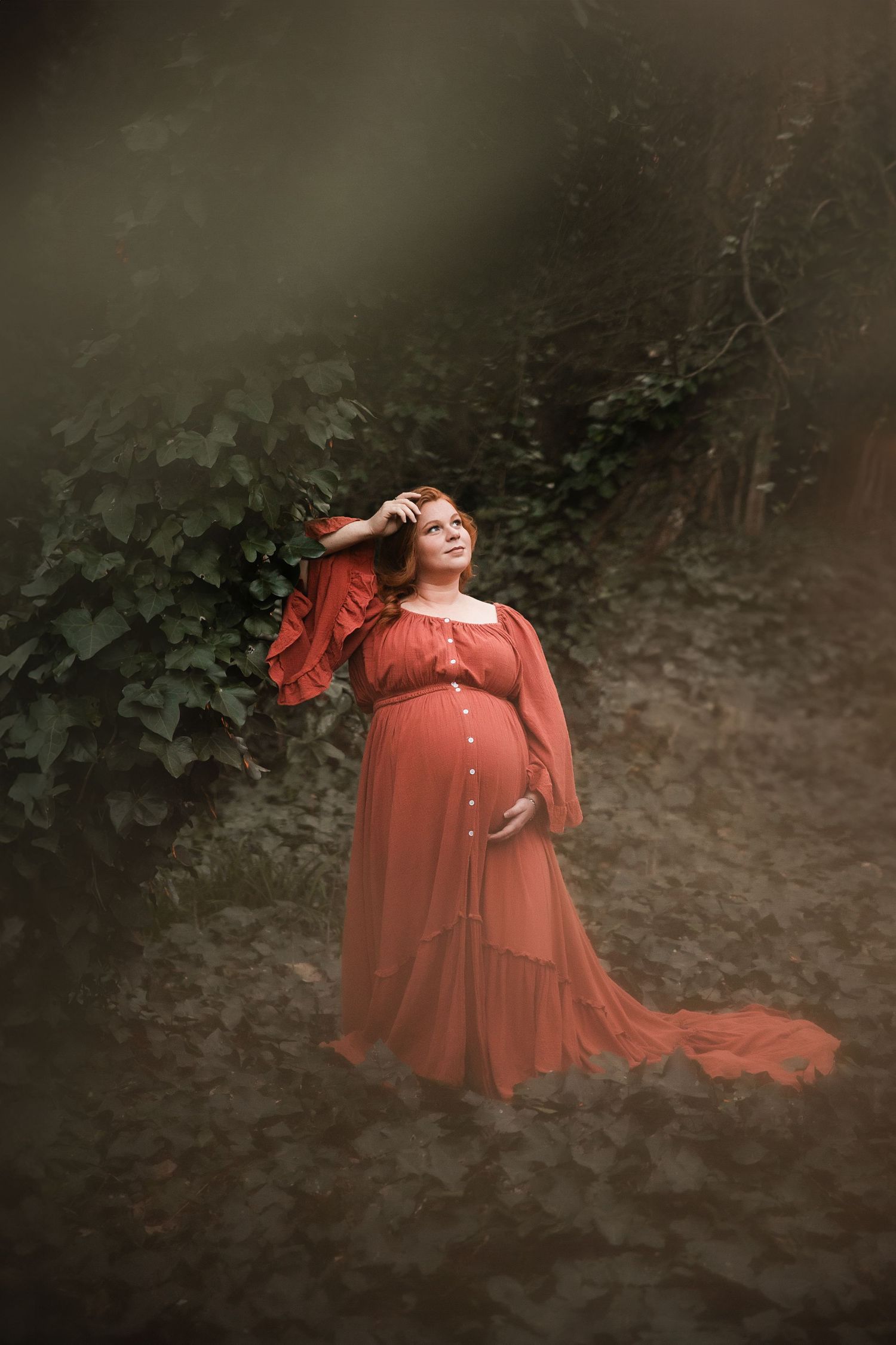 Pregnant woman in flowing rust-orange gown poses gracefully outdoors among lush ivy, one hand in her red hair.
