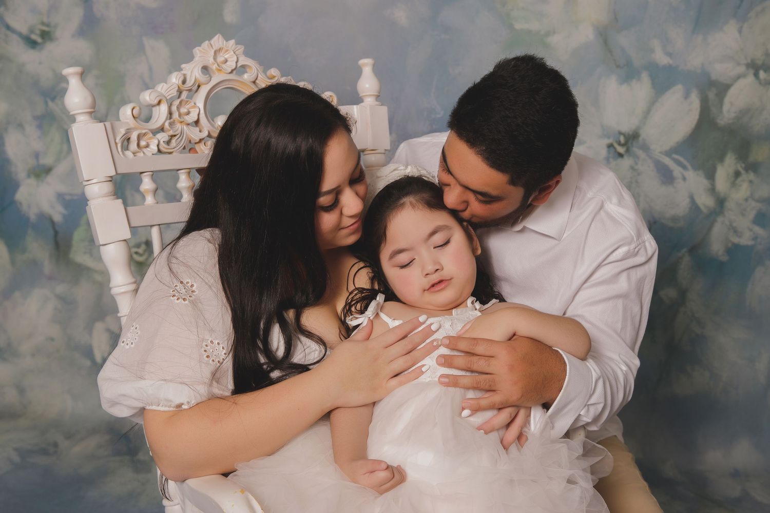 Three family members in white outfits share an intimate embrace against a soft floral backdrop.