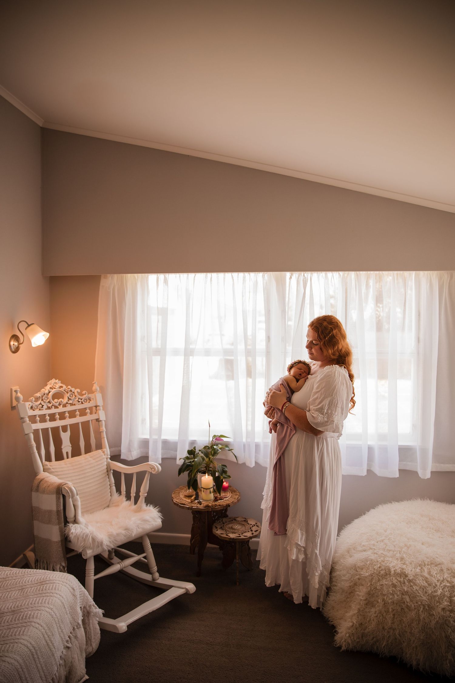 A mother in a white dress holds her newborn baby by a bright window in a cozy nursery with a rocking chair.