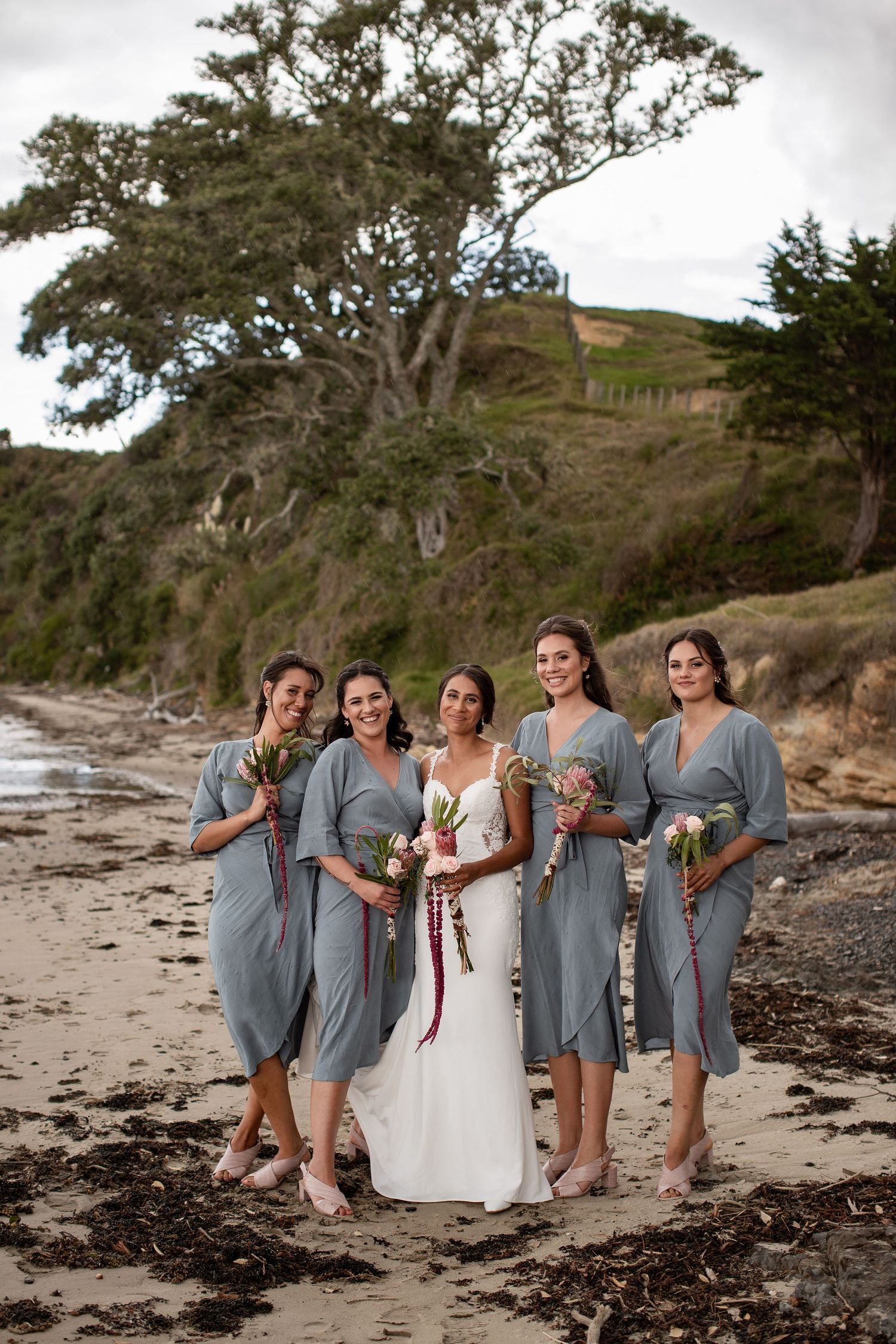 A wedding party in gray dresses poses together on a rocky beach with trees in the background.