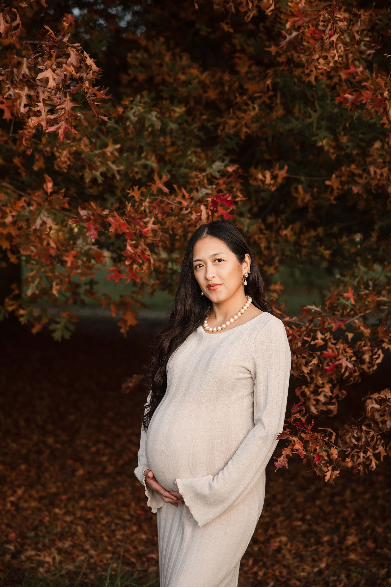 Pregnant woman in beige dress holding belly, standing before vibrant autumn red foliage in a serene outdoor setting.