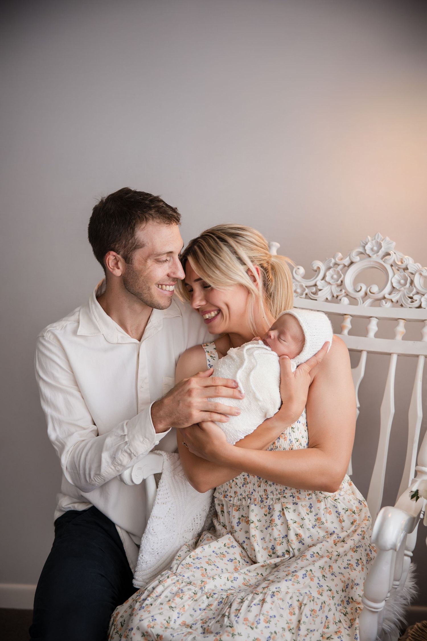 Happy couple smiling at their newborn baby, sitting on a white ornate chair in a bright, cozy room.
