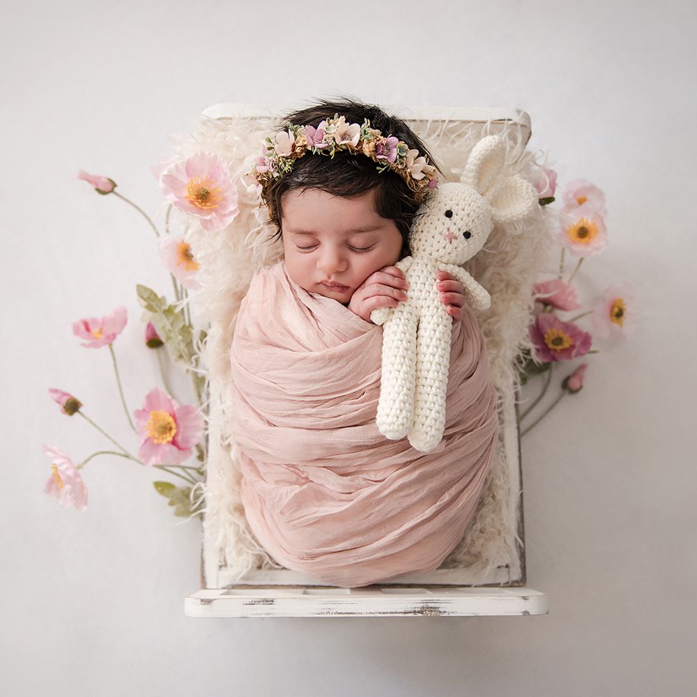 Newborn baby girl swaddled in pink wrap, wearing floral crown, sleeping with knit bunny toy in white wooden bed with pink flowers.