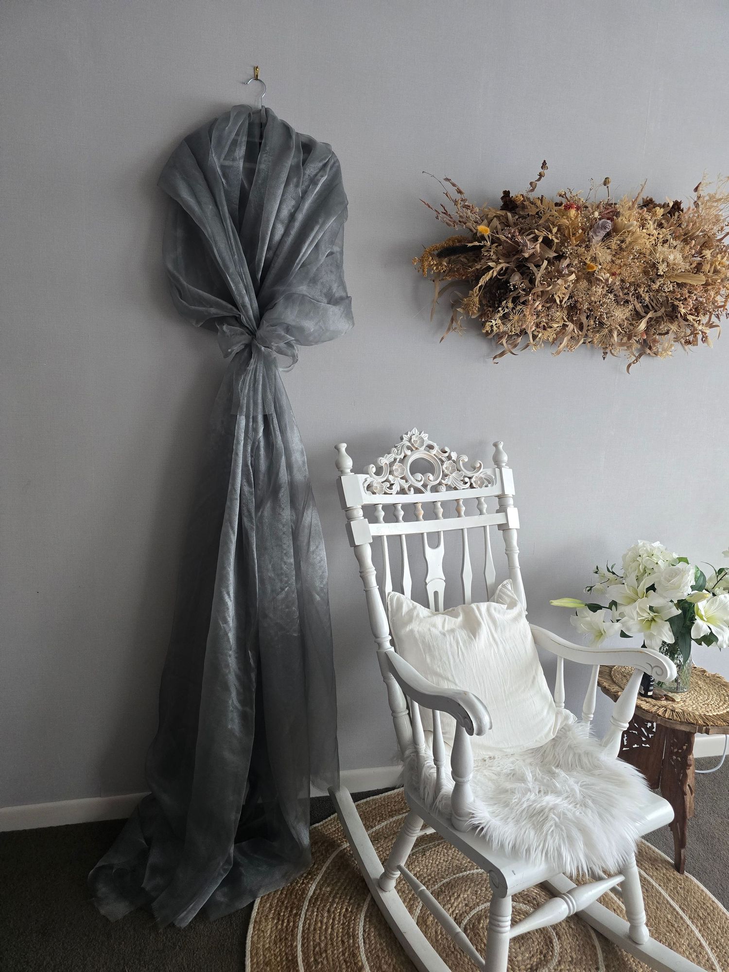 Elegant white rocking chair with floral decor, gray tulle drape, and dried flower wreath in a cozy room.