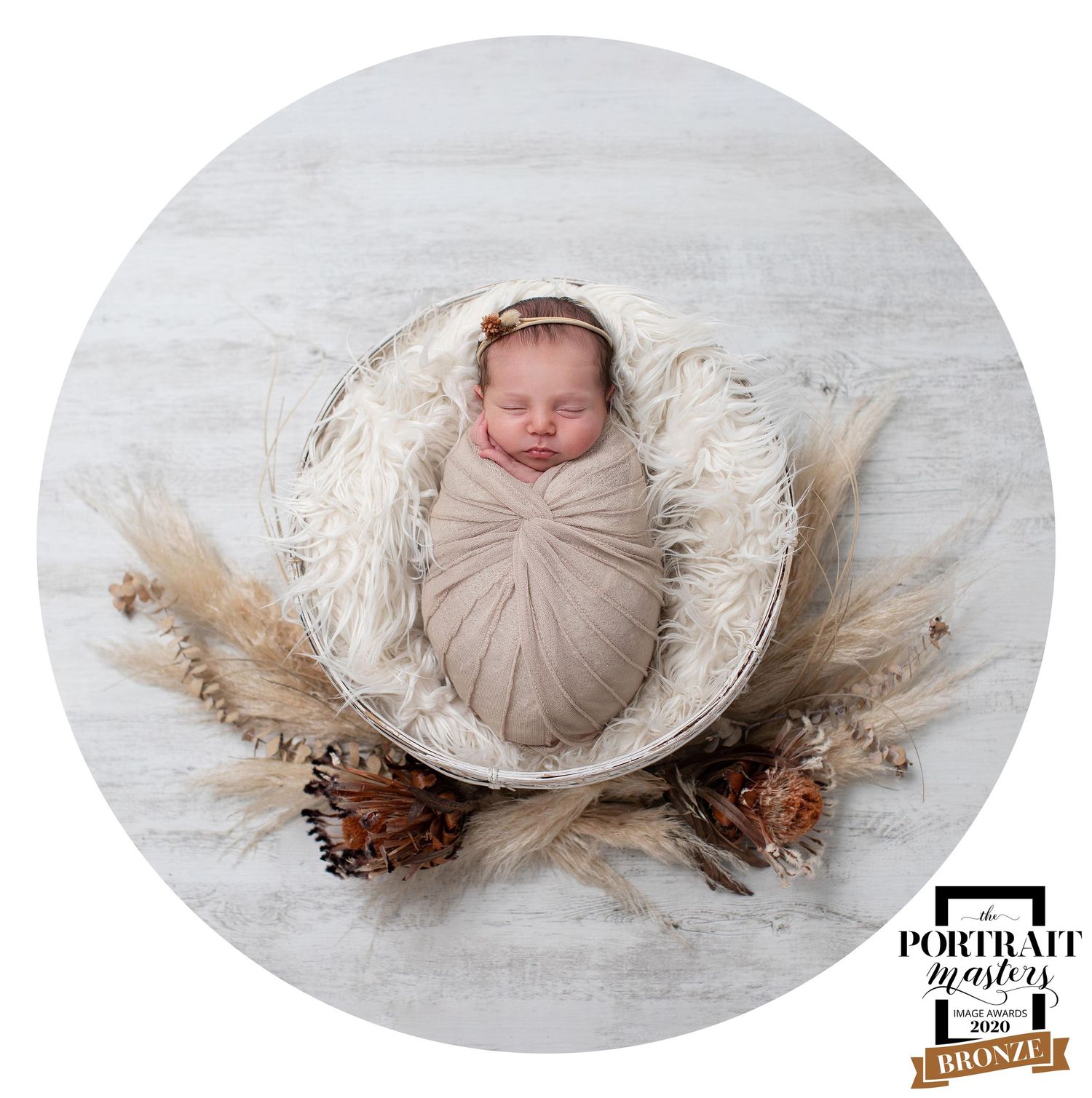 Newborn baby swaddled in beige fabric lying in oval-shaped basket with dried floral decorations on white backdrop.