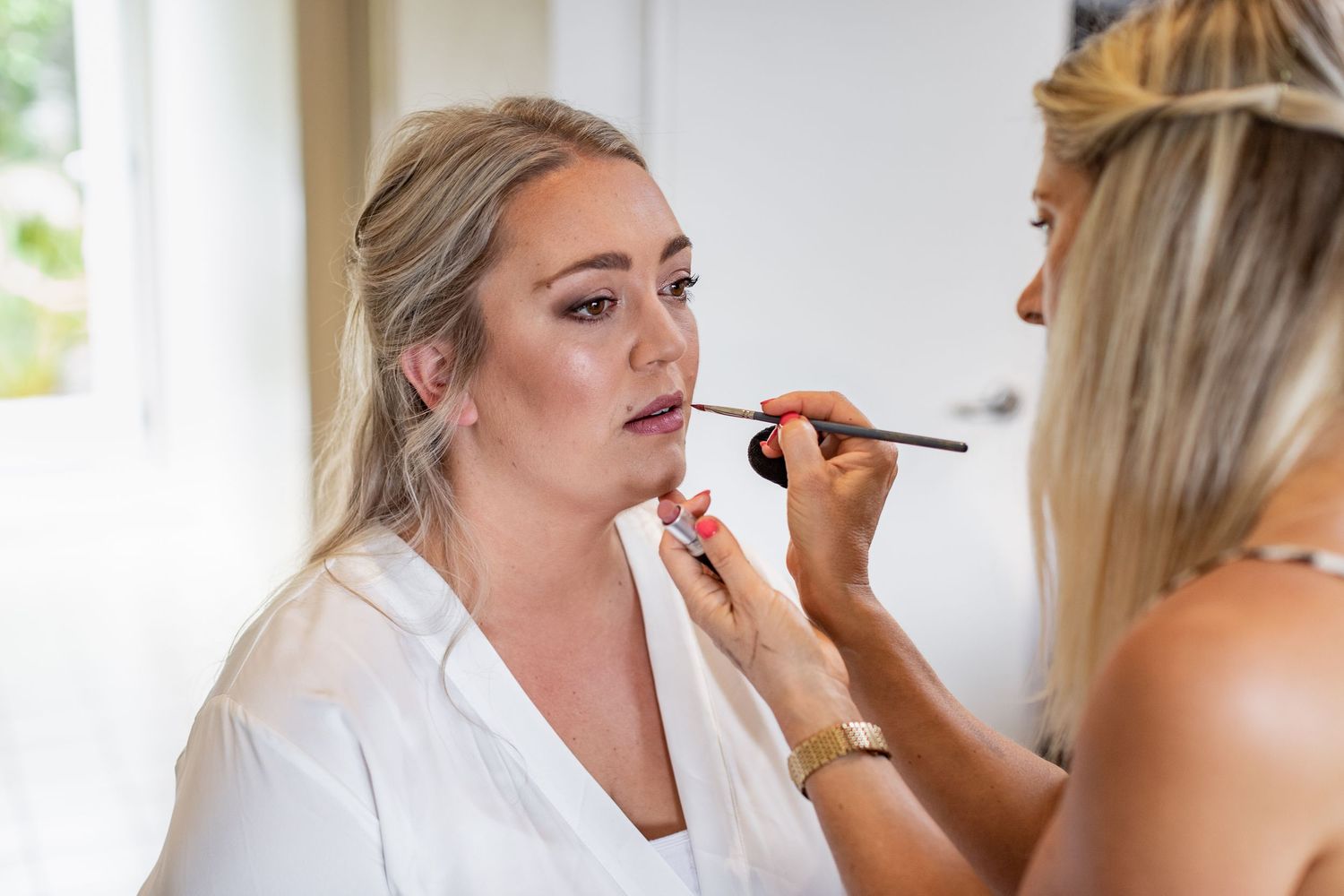 A makeup artist applies lipstick on someone wearing a white robe during wedding preparations.
