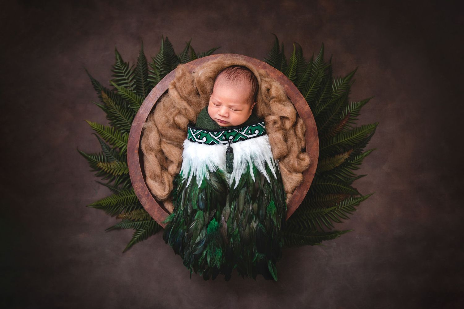 Sleeping newborn in wooden bowl surrounded by green fern leaves and brown fabric in artistic portrait setting. Cambridge Waikato NZ Photogra