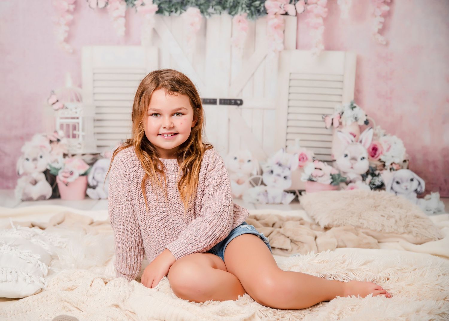 A young person sits on plush bedding in a pink sweater against a floral backdrop with white shutters.