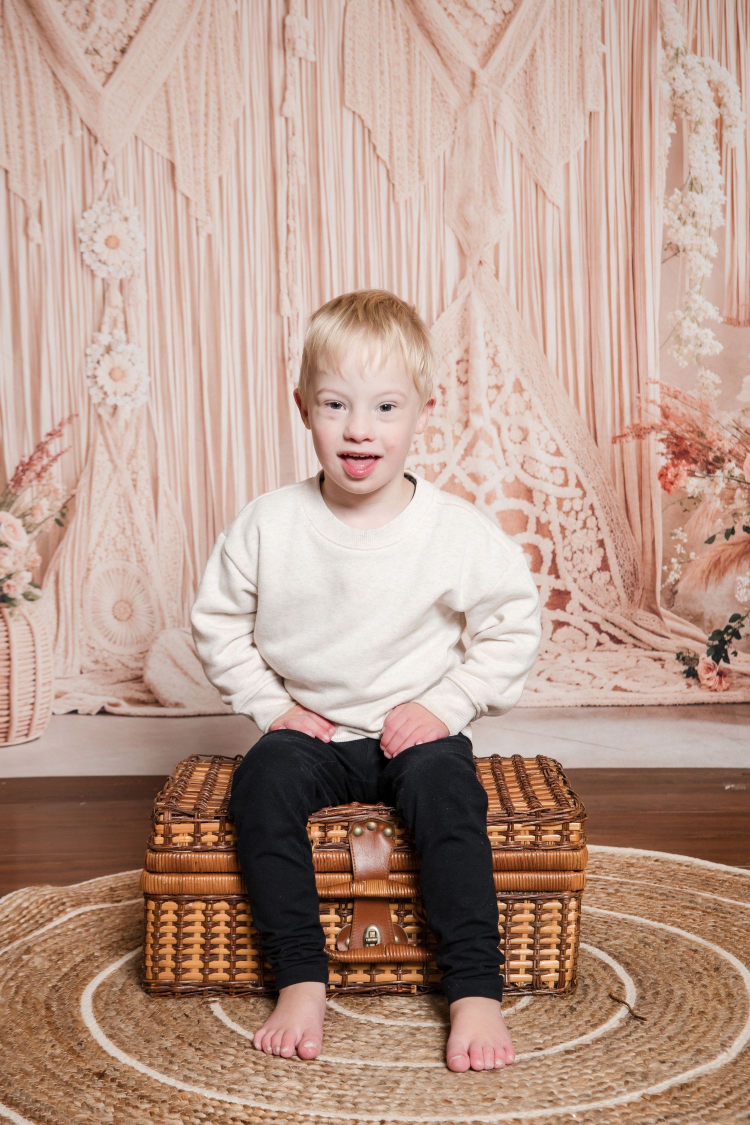 A child sits on a brown wicker suitcase against a pink floral backdrop wearing a white sweater and dark pants.