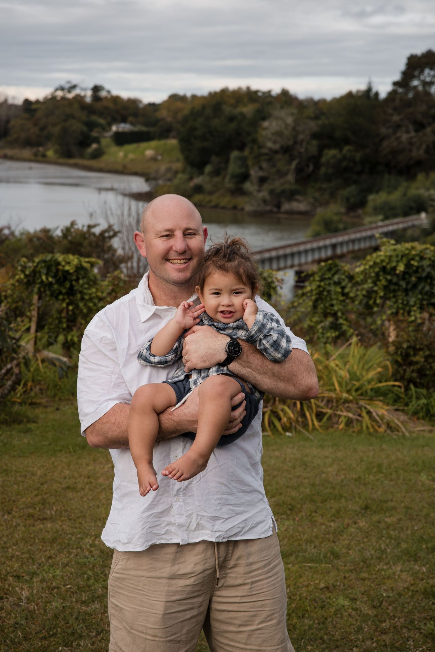 Smiling bald man holding a happy toddler outdoors near a river bridge surrounded by lush greenery.