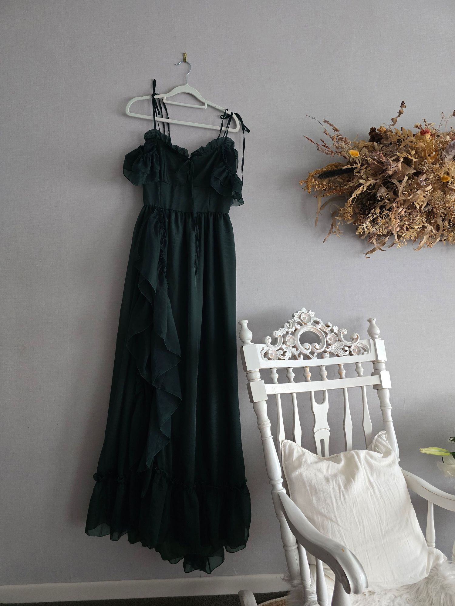 Dark green chiffon ruffle maxi dress on hanger displayed against gray wall near white vintage chair.