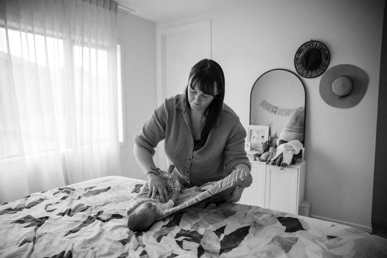 Black and white photo of a mother tenderly wrapping her newborn baby on a bed at home.