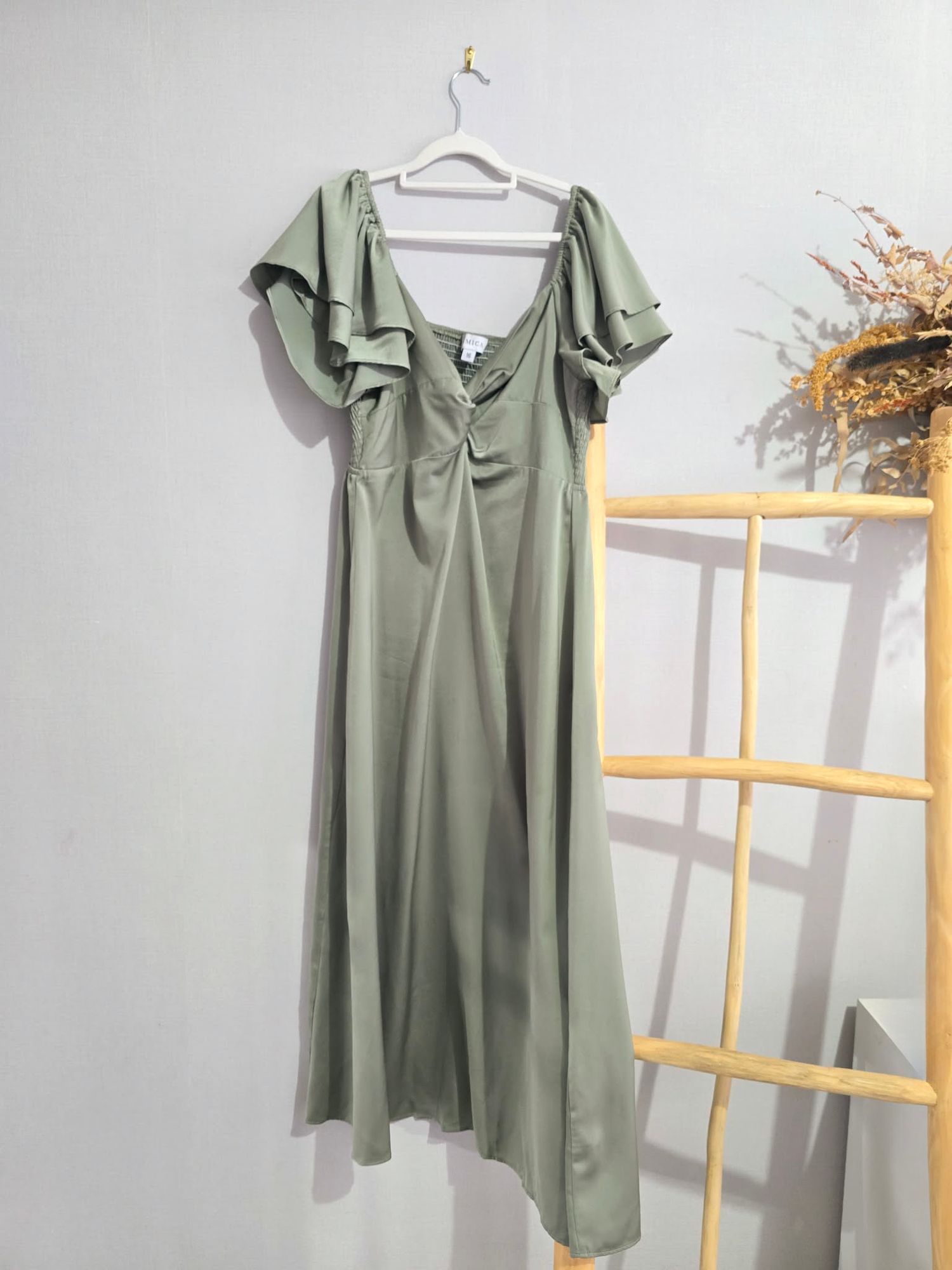 Sage green satin midi dress with flutter sleeves and deep V-neckline, hanging on a hanger against a white wall.