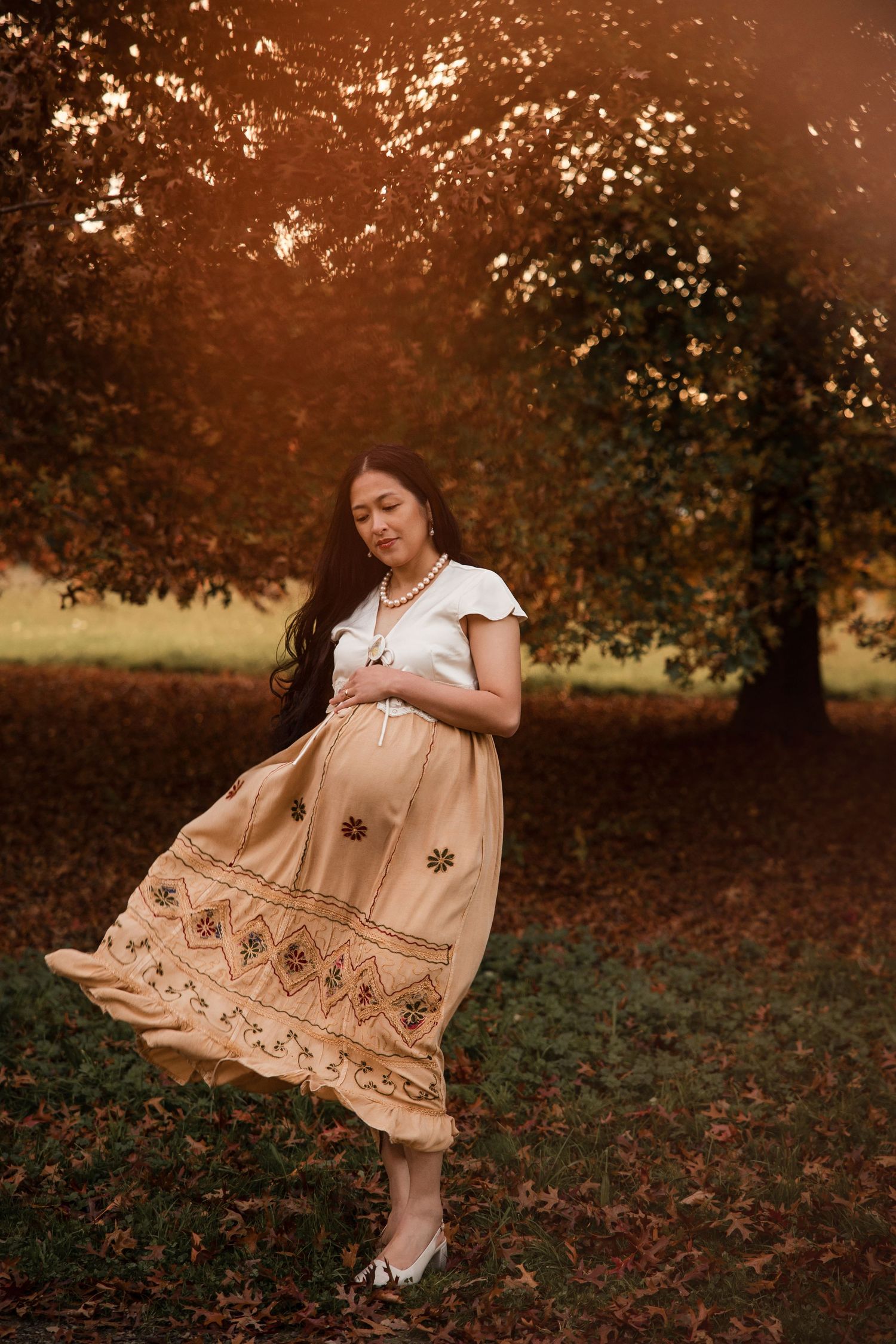 Pregnant woman in beige embroidered skirt stands under autumn tree at golden sunset in a serene outdoor setting.
