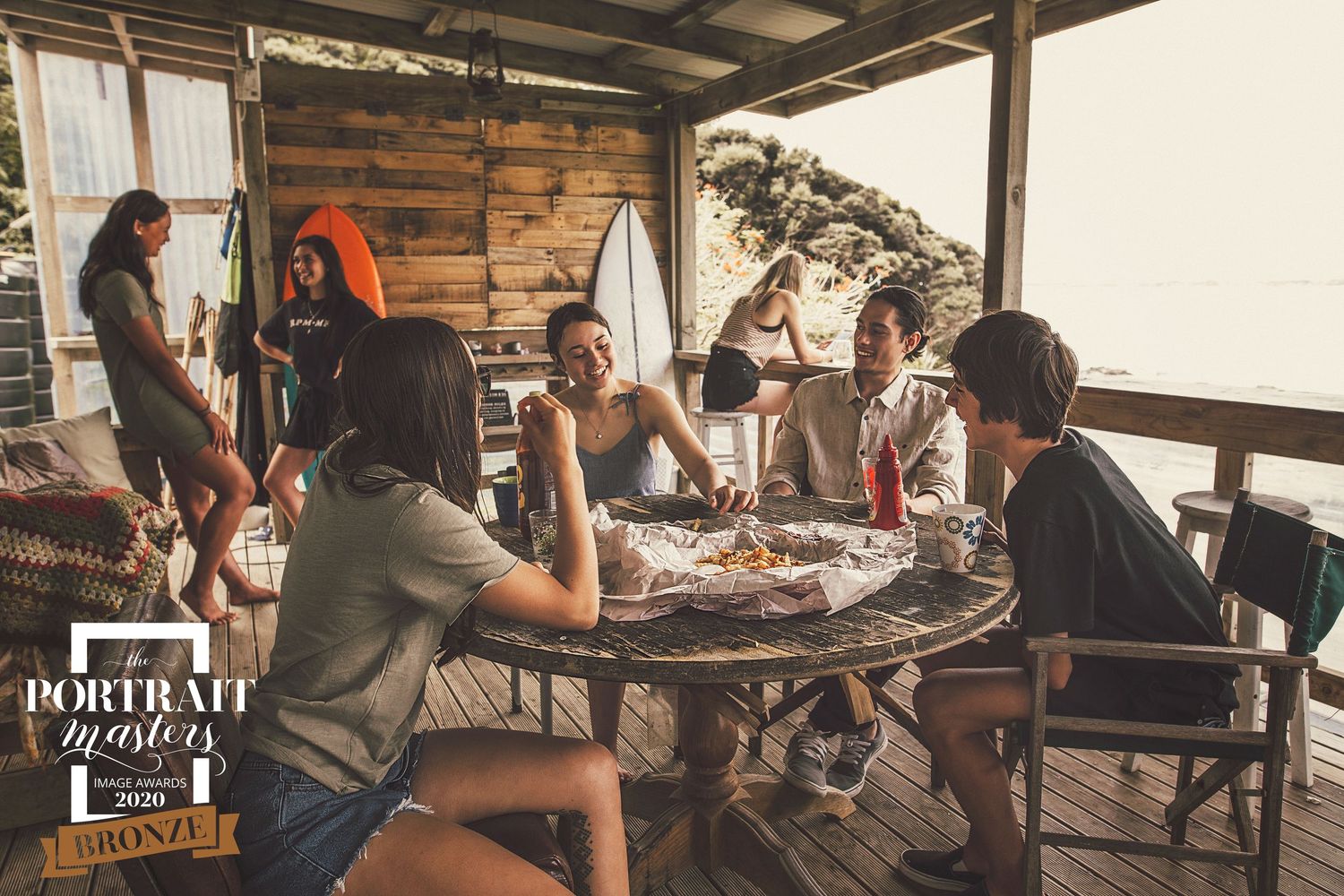 A group gathers around a wooden table on a covered porch for casual drinks and conversation in warm lighting.
