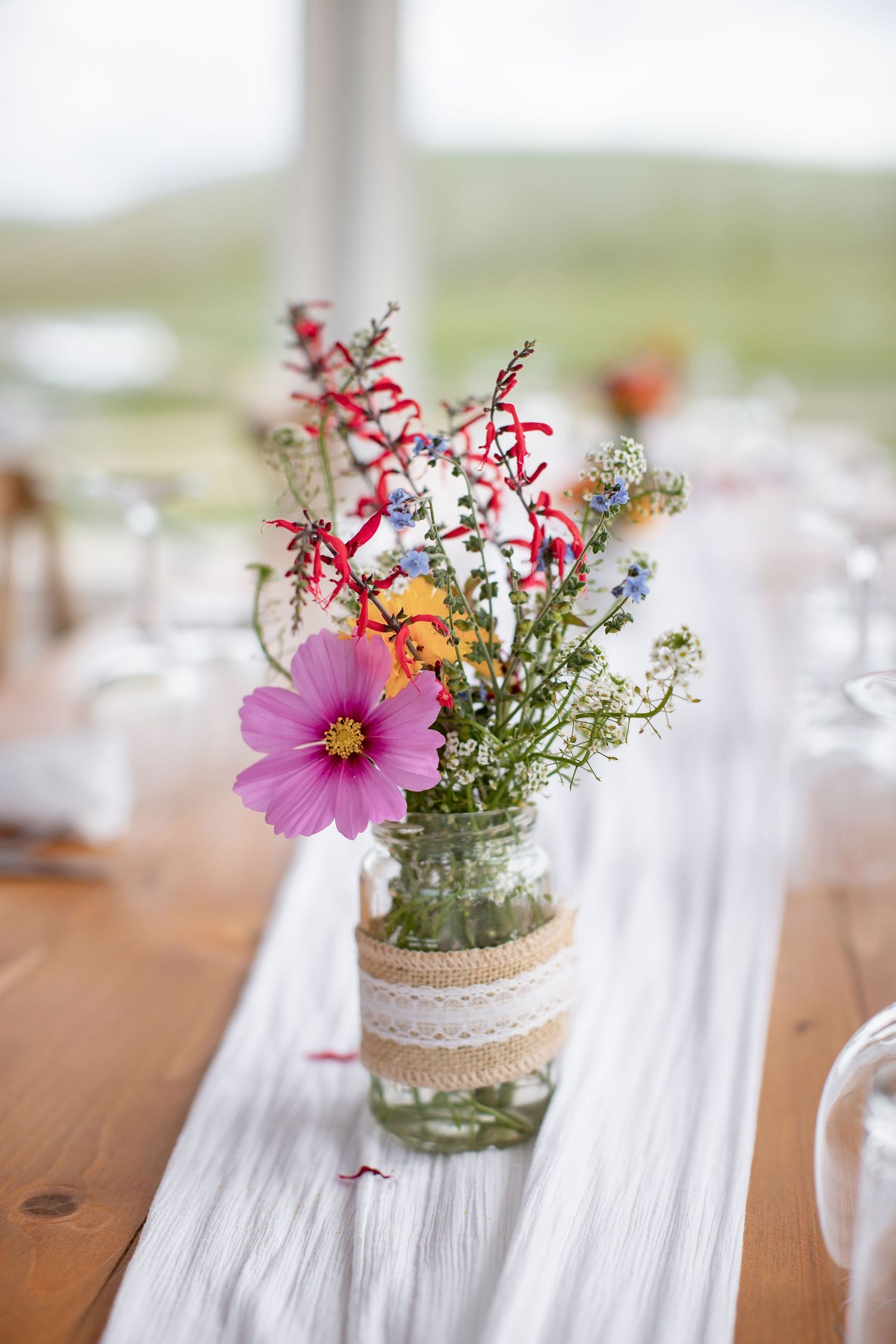 Rustic wedding centerpieces with pink flowers and greenery in mason jars wrapped with lace on a wooden table.