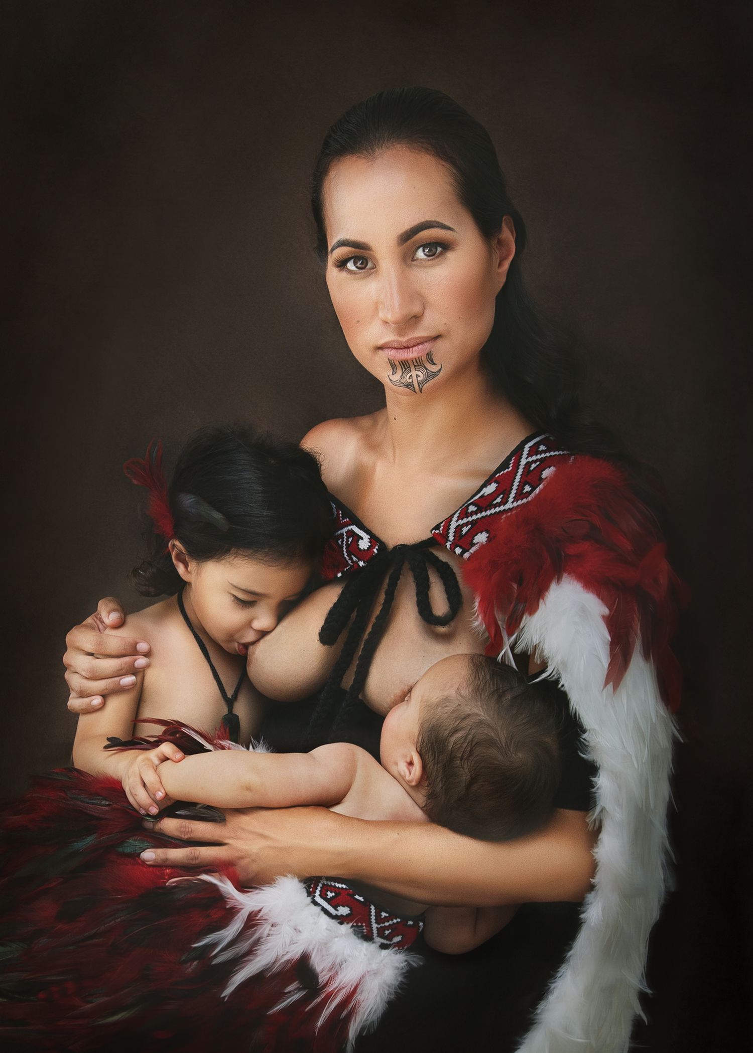Maori woman in traditional red and white feathered cloak breastfeeding two children, showcasing cultural heritage.