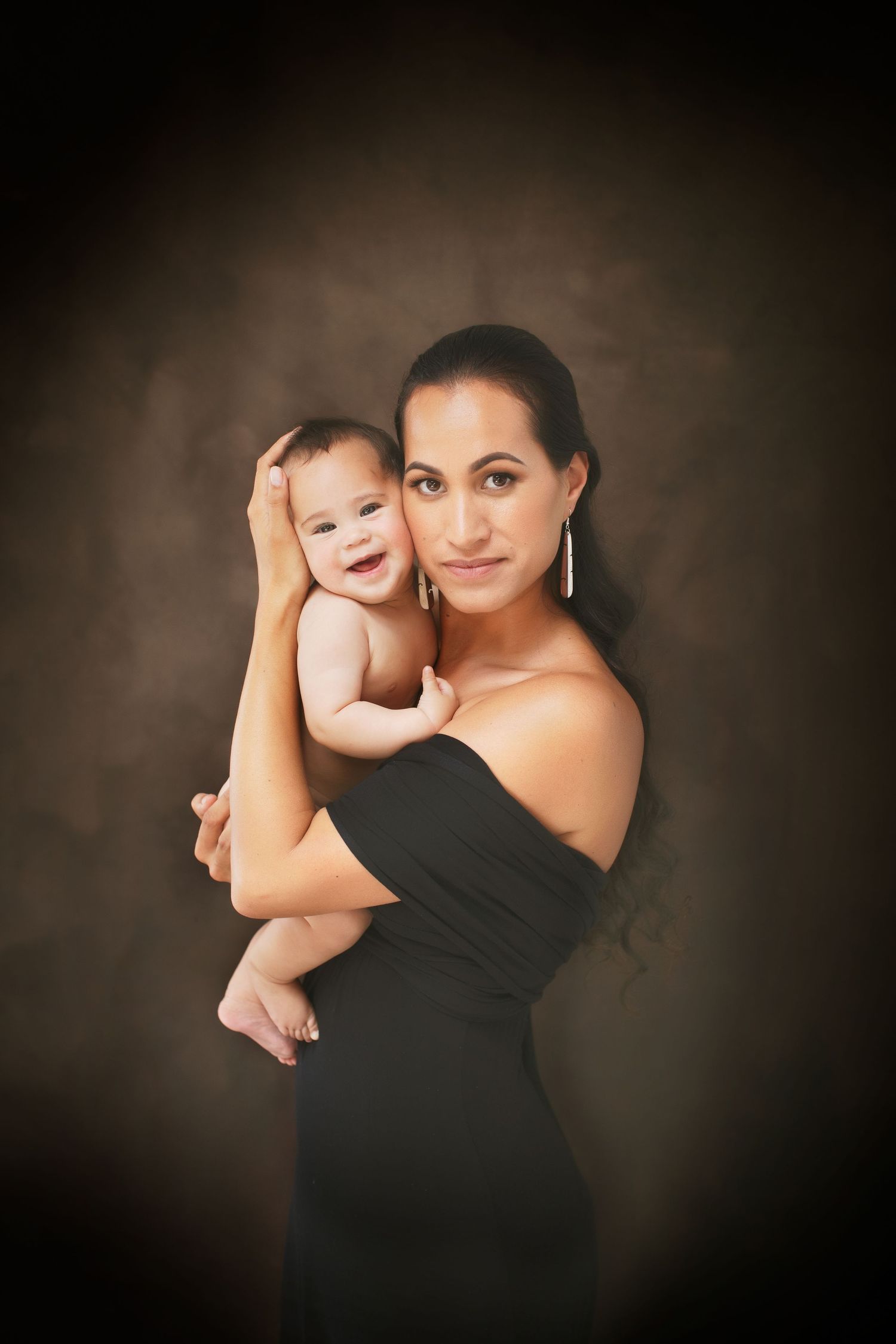 Mother in black dress holds smiling baby in intimate studio portrait.