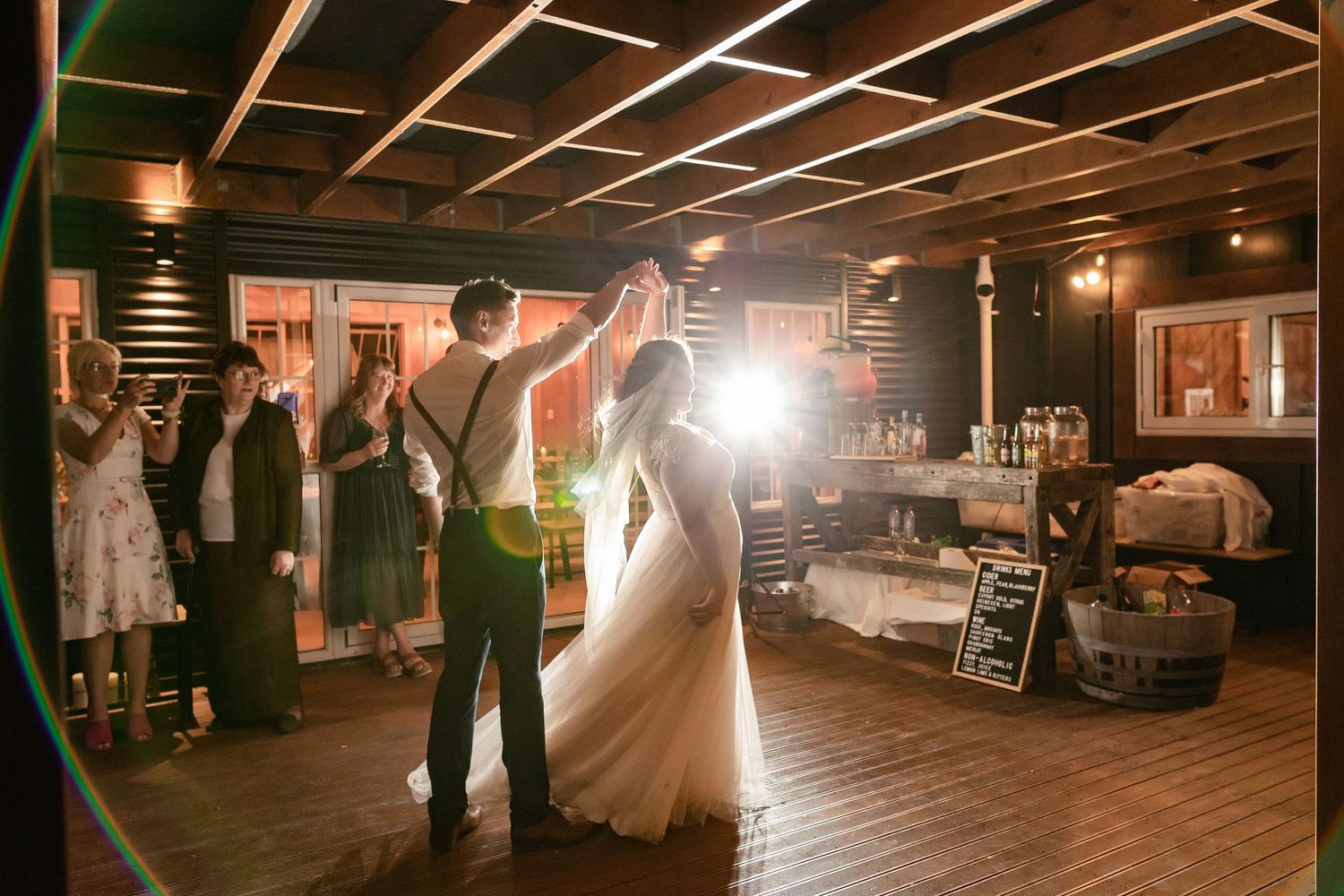 Wedding reception guests dance in a dark rustic barn venue illuminated by warm string lights and lanterns.
