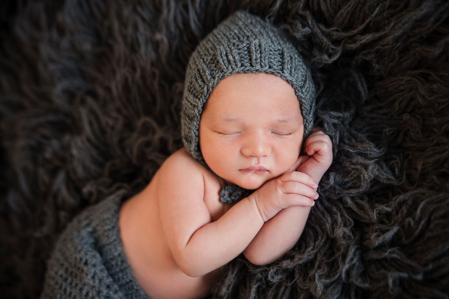 Sleeping newborn baby wearing gray knit hat and laying on dark furry blanket with hands tucked under chin.
