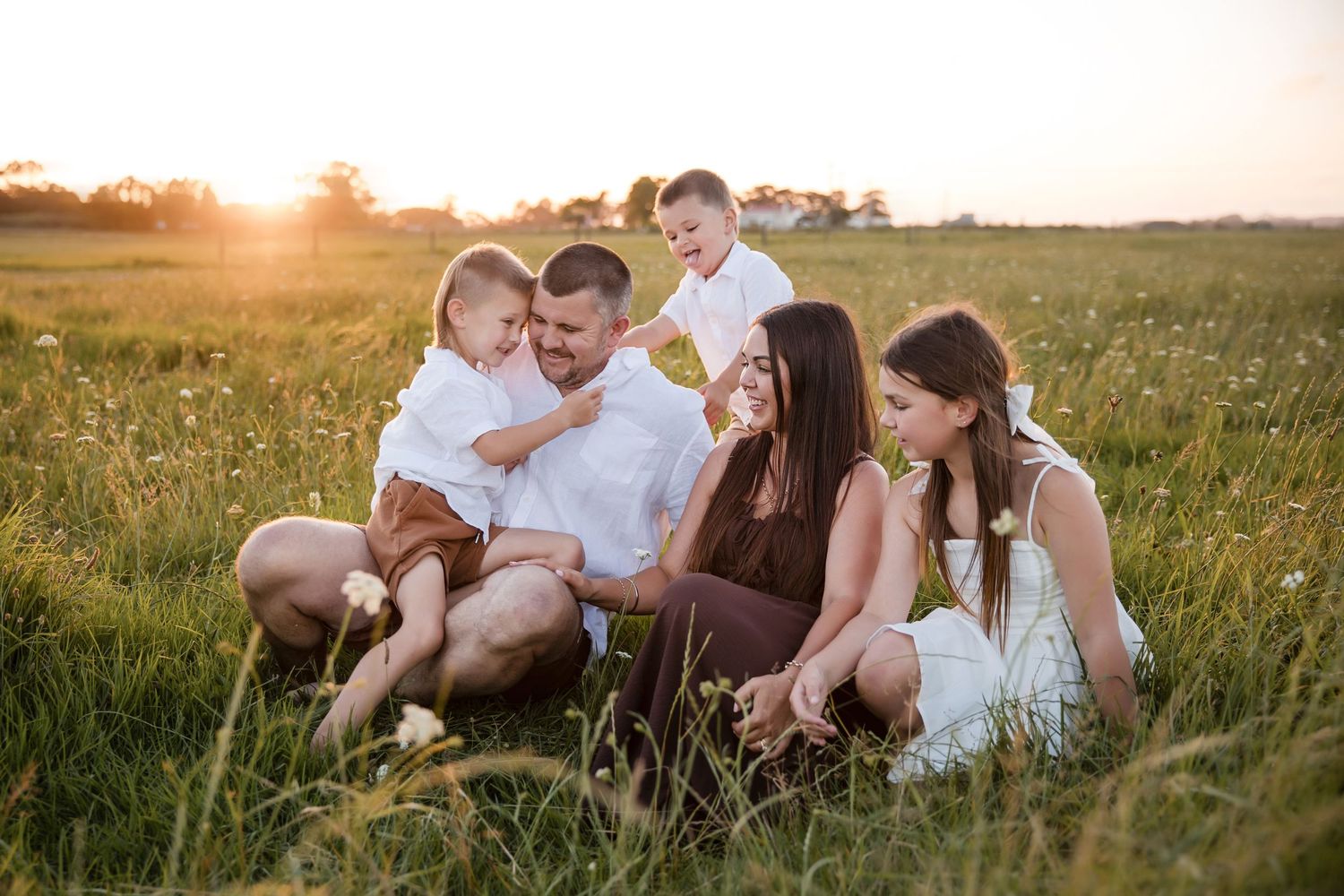 Family of four sitting in a sunlit green field at golden hour, smiling and enjoying quality time together.