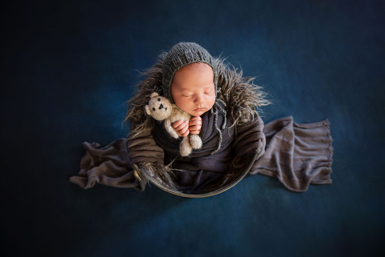 Newborn baby sleeping in a bowl with a teddy bear, wearing a knit hat on a blue background.