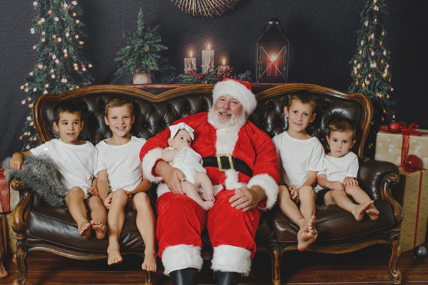 A festive holiday scene with children and Santa Claus sitting on a vintage brown couch surrounded by decorated Christmas trees.