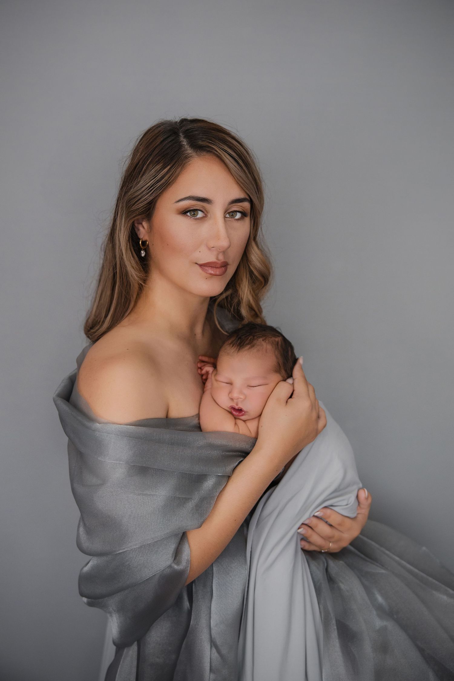 A young mother wrapped in a gray silk cloth tenderly holds her sleeping newborn baby against a gray background.