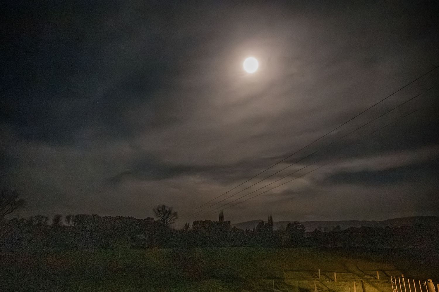 A bright full moon illuminates dark clouds over a silhouetted rural landscape at night.
