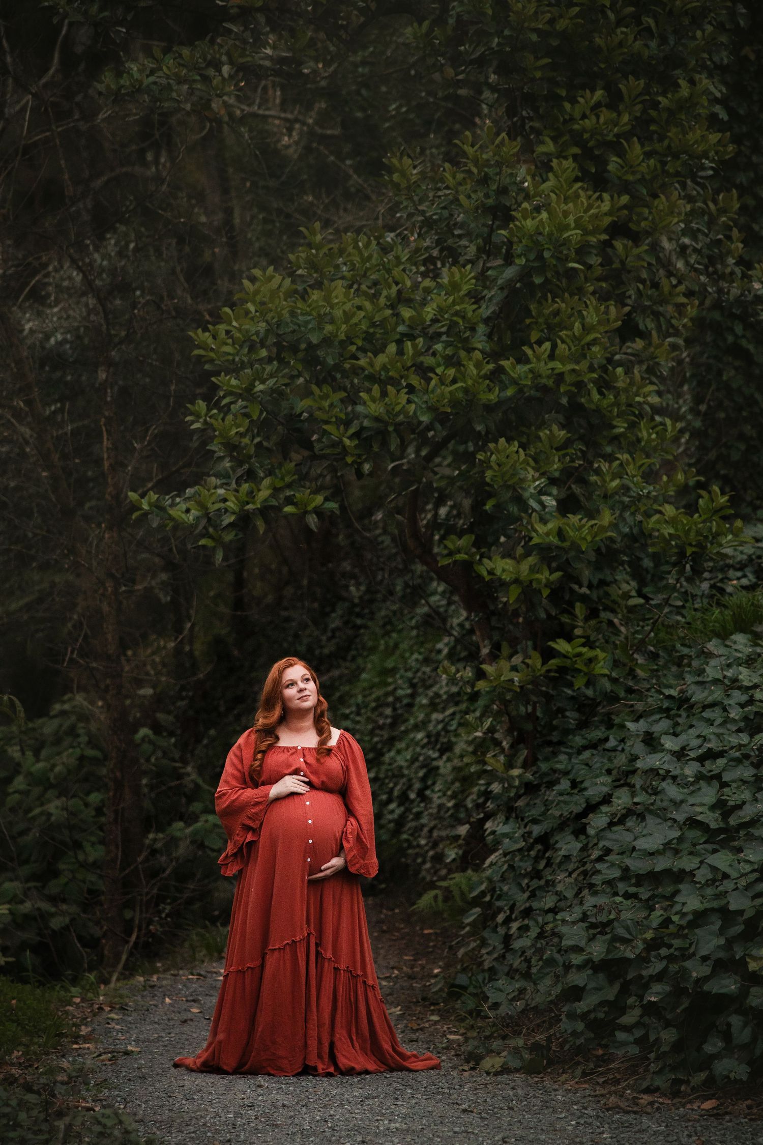 A person in a flowing rust-colored dress poses in a forest setting against dark green foliage background.