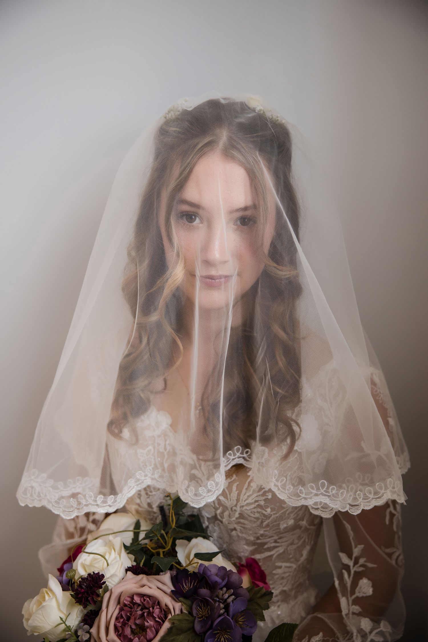 A bride in a lace-trimmed veil holds a romantic bouquet of pink and purple flowers against a soft gray background.