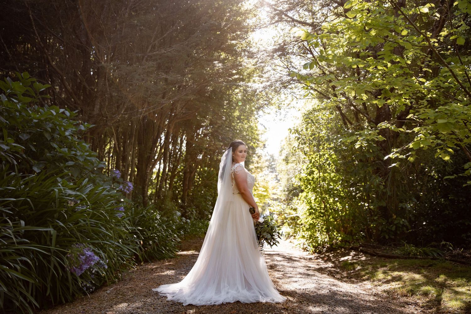 A bride in a flowing white wedding dress stands on a sunlit forest path surrounded by lush greenery.