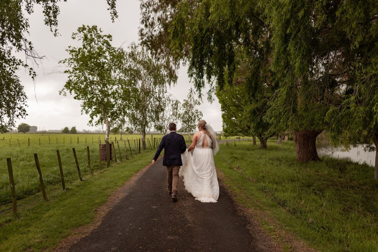 Two people walk along a gravel path between willow trees and a fence on a green country estate.
