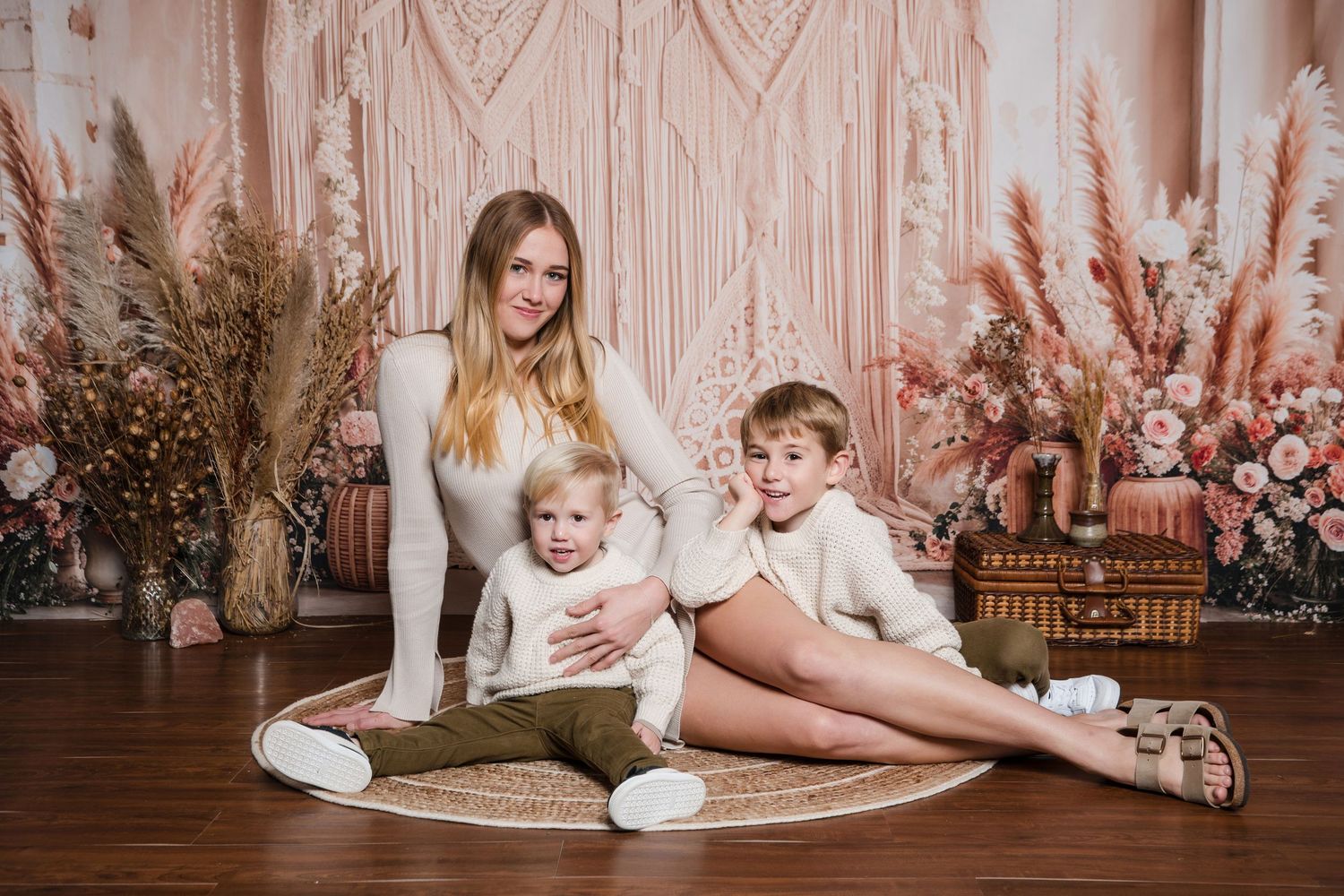 A family photo session featuring pampas grass and floral decorations against a blush pink macrame backdrop on hardwood floor.
