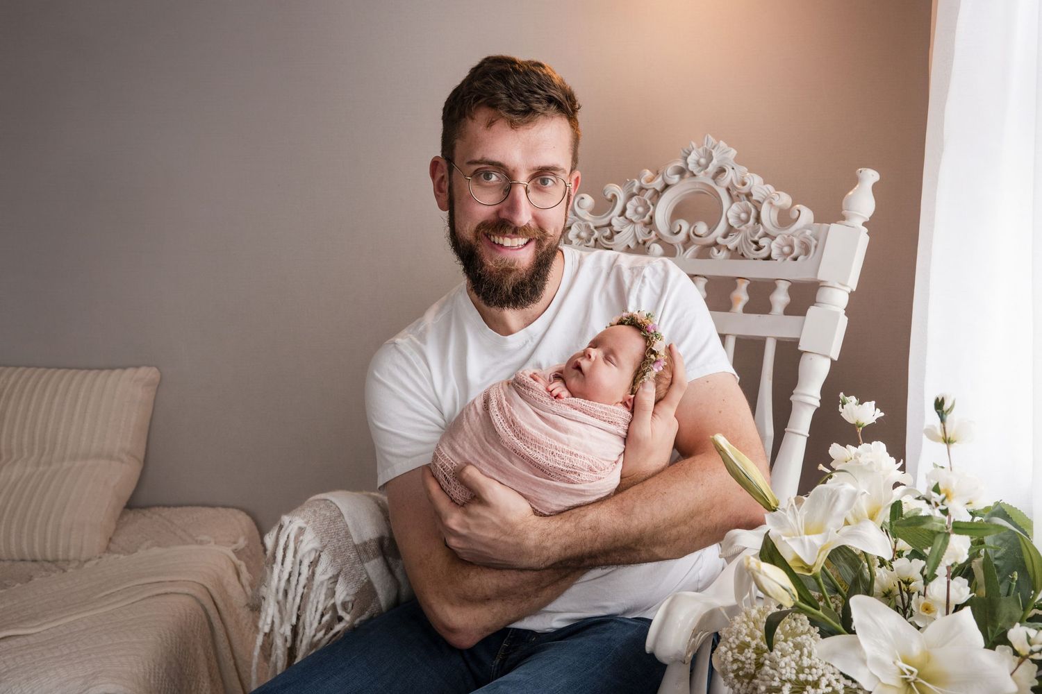 A person in a white shirt holds a swaddled newborn baby while sitting in an ornate white chair near floral arrangements.
