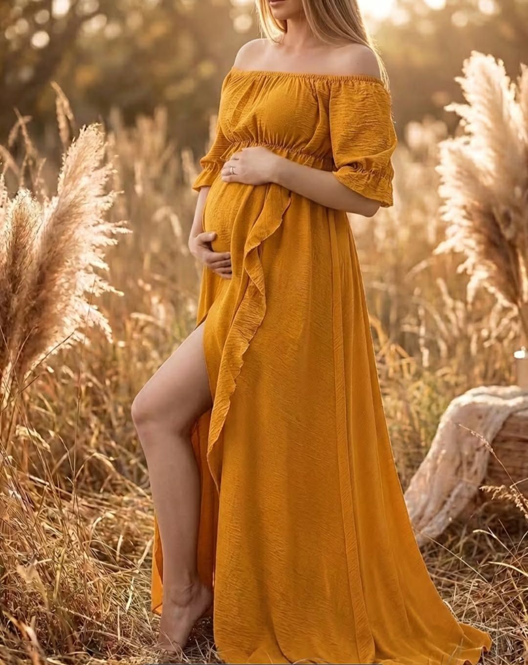 Pregnant woman in golden yellow off-shoulder maxi dress with ruffle slit, posing outdoors among tall golden grasses.