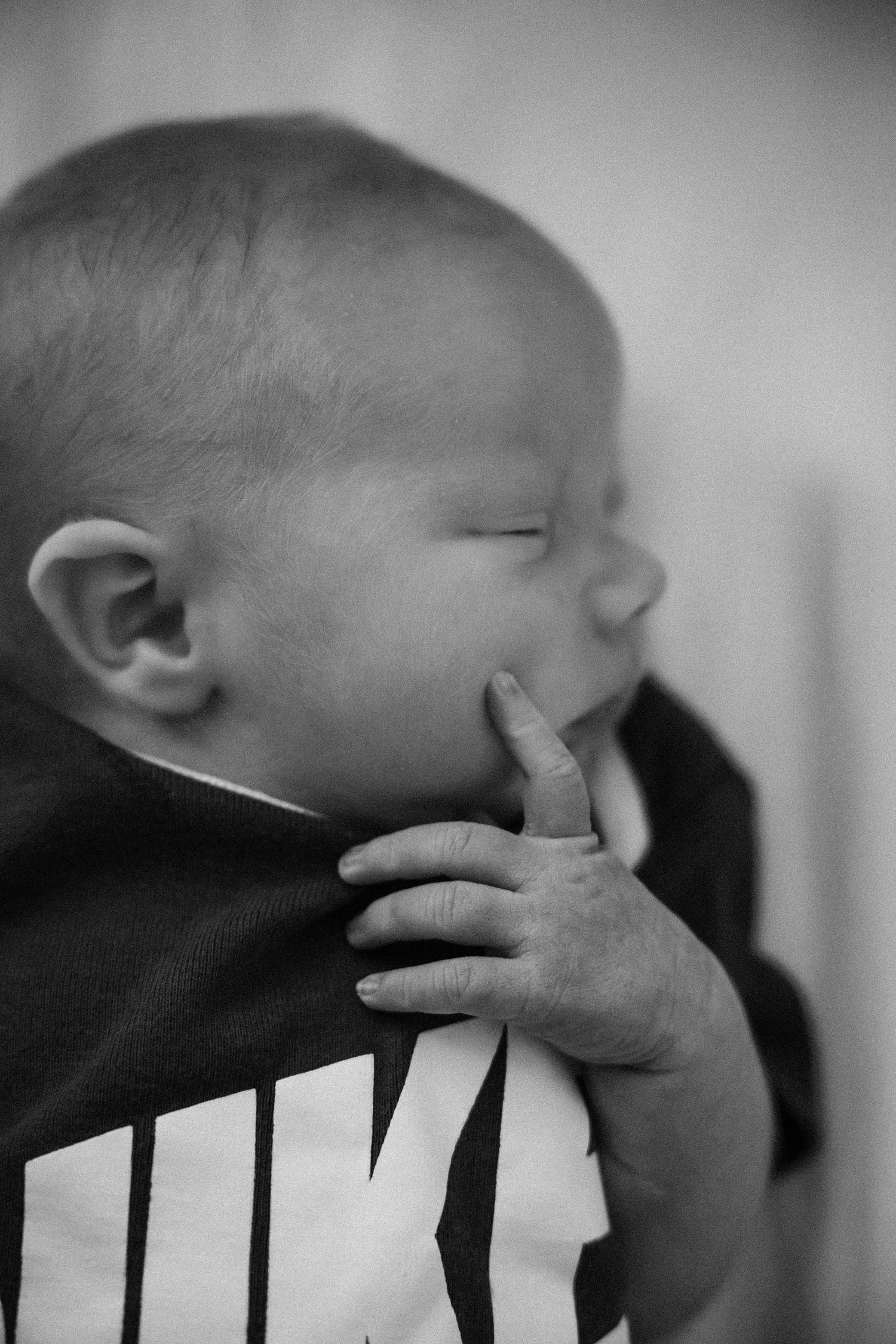 Black and white photo of a newborn baby in a Nike onesie, sucking their finger with eyes closed.